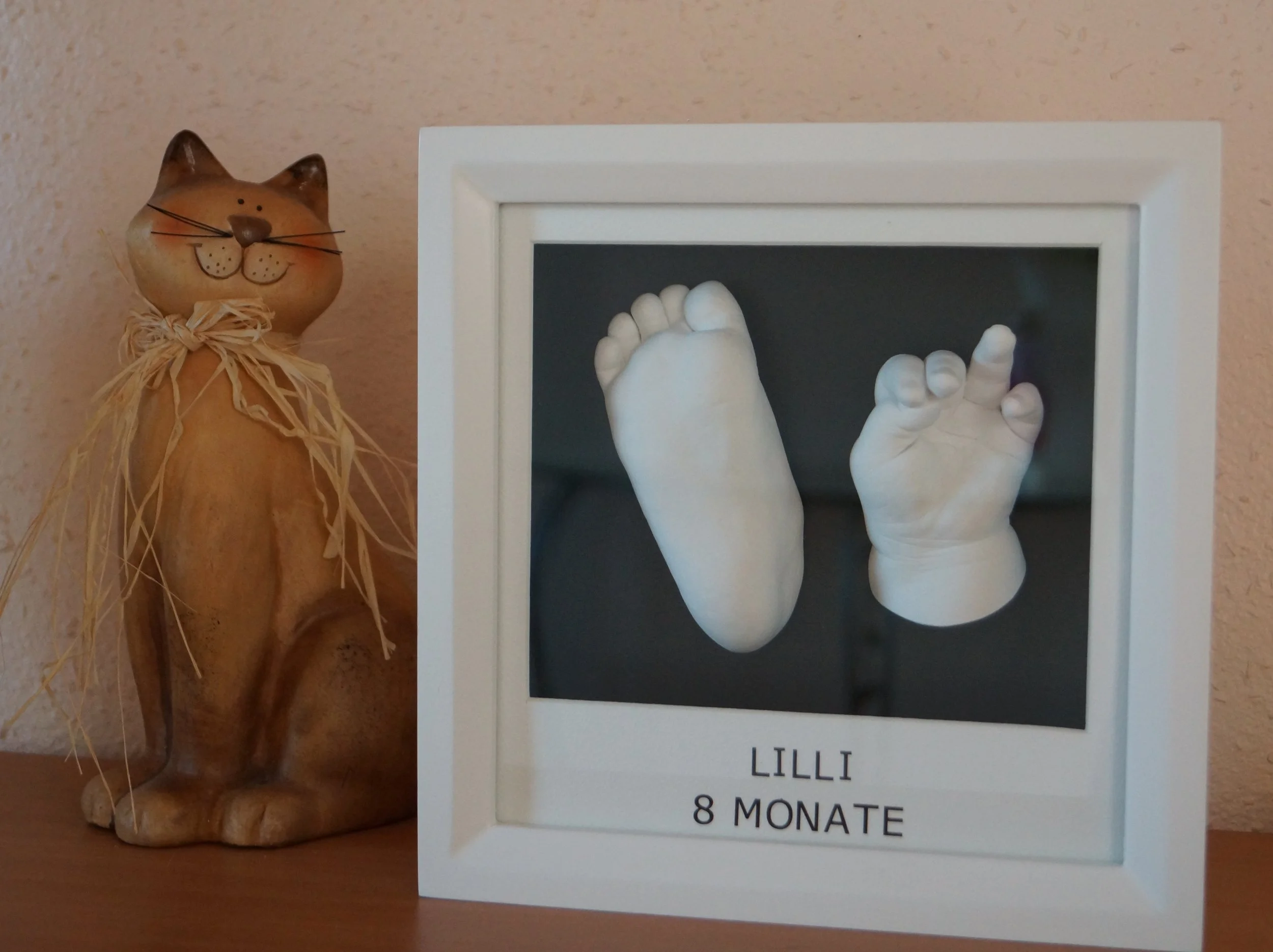 Eine Figur eines Katzenbabys aus Keramik neben einem kleinen weißen Bilderrahmen mit Babyfüßen darin beim Fotografieren.