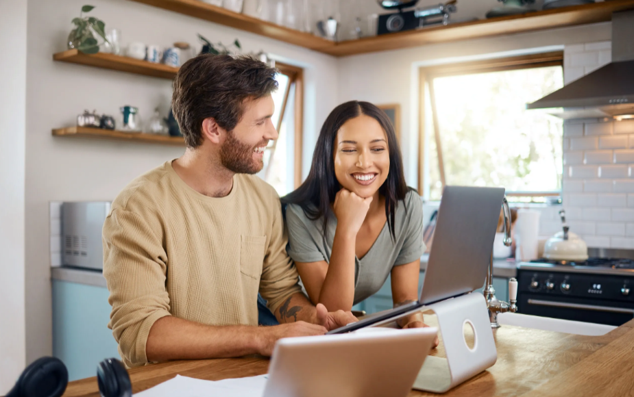 A smiling man and woman sitting at a kitchen counter, looking at a laptop, with a bright kitchen background.