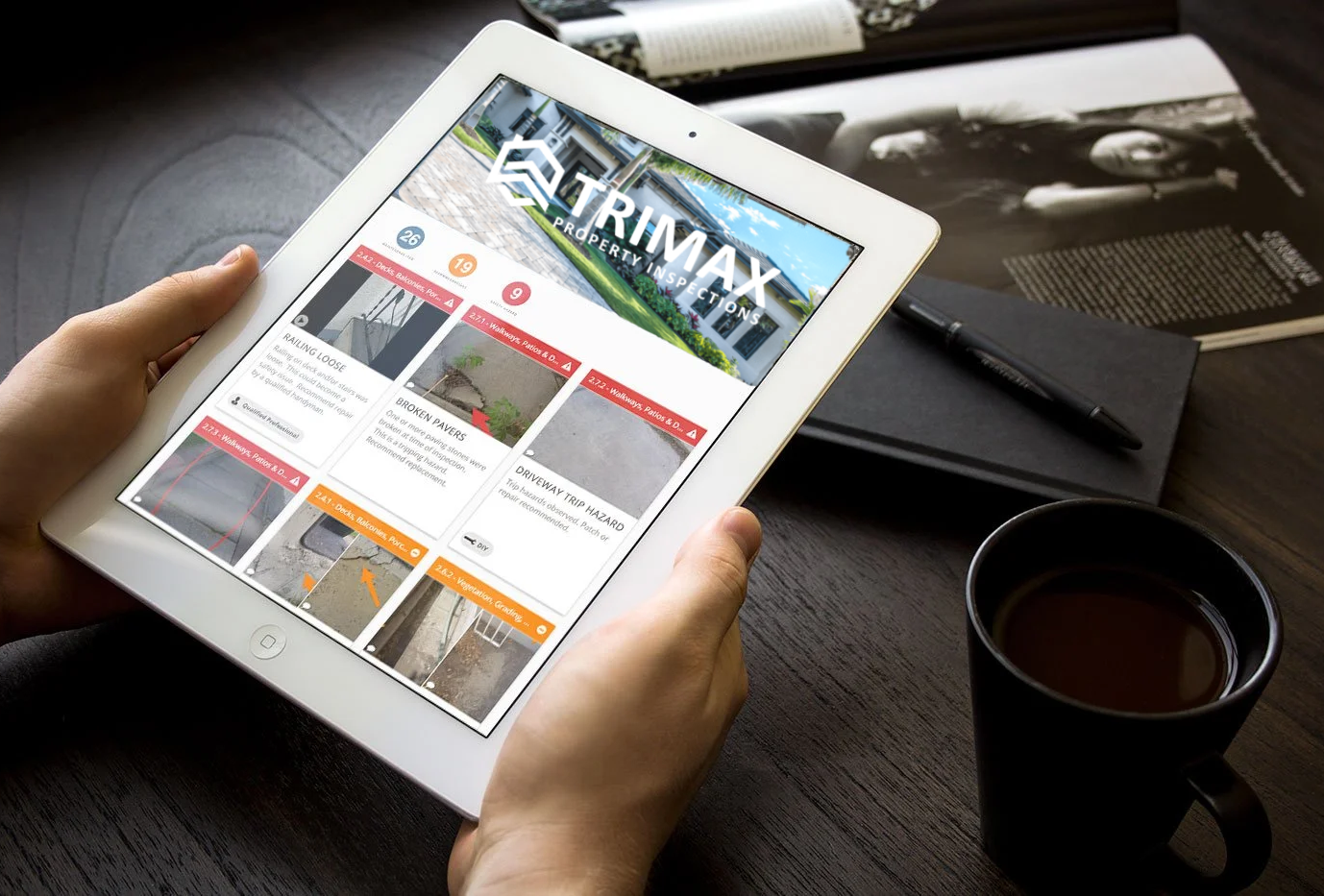 Person holding a white tablet displaying a property inspection app, sitting at a dark wooden table with a black notebook, pen, and a cup of coffee nearby.