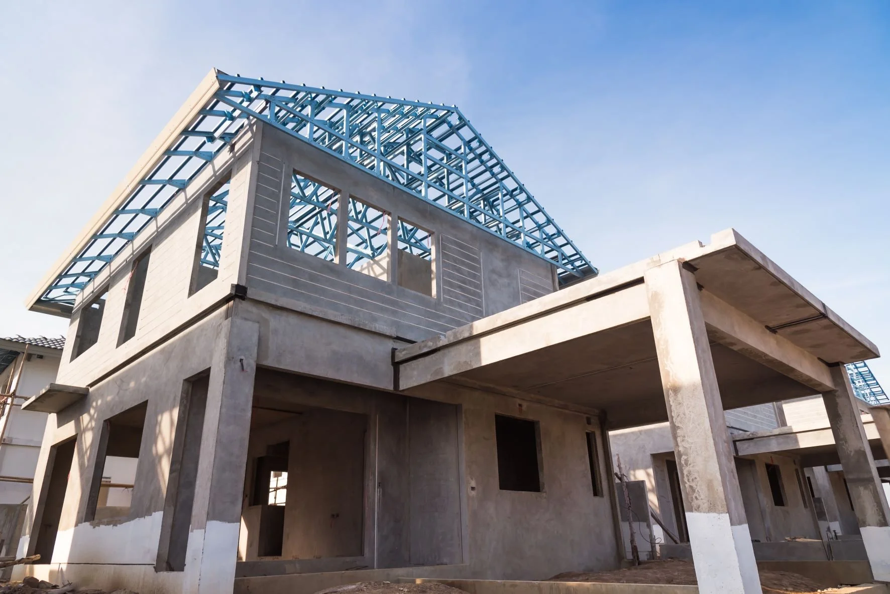 An under-construction two-story house with a concrete structure and a blue metal roof framework. The house features large windows and open doorways, and it is set against a clear sky.