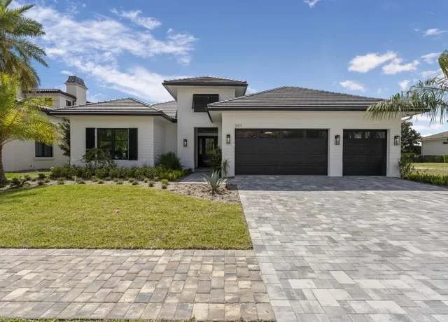 Modern white house with black garage doors, a paved driveway, manicured lawn, and palm trees under a partly cloudy sky.