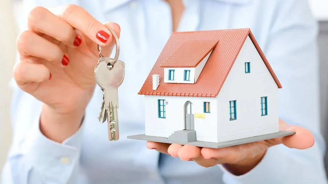 Person holding a small model house in one hand and a key with a keyring in the other.