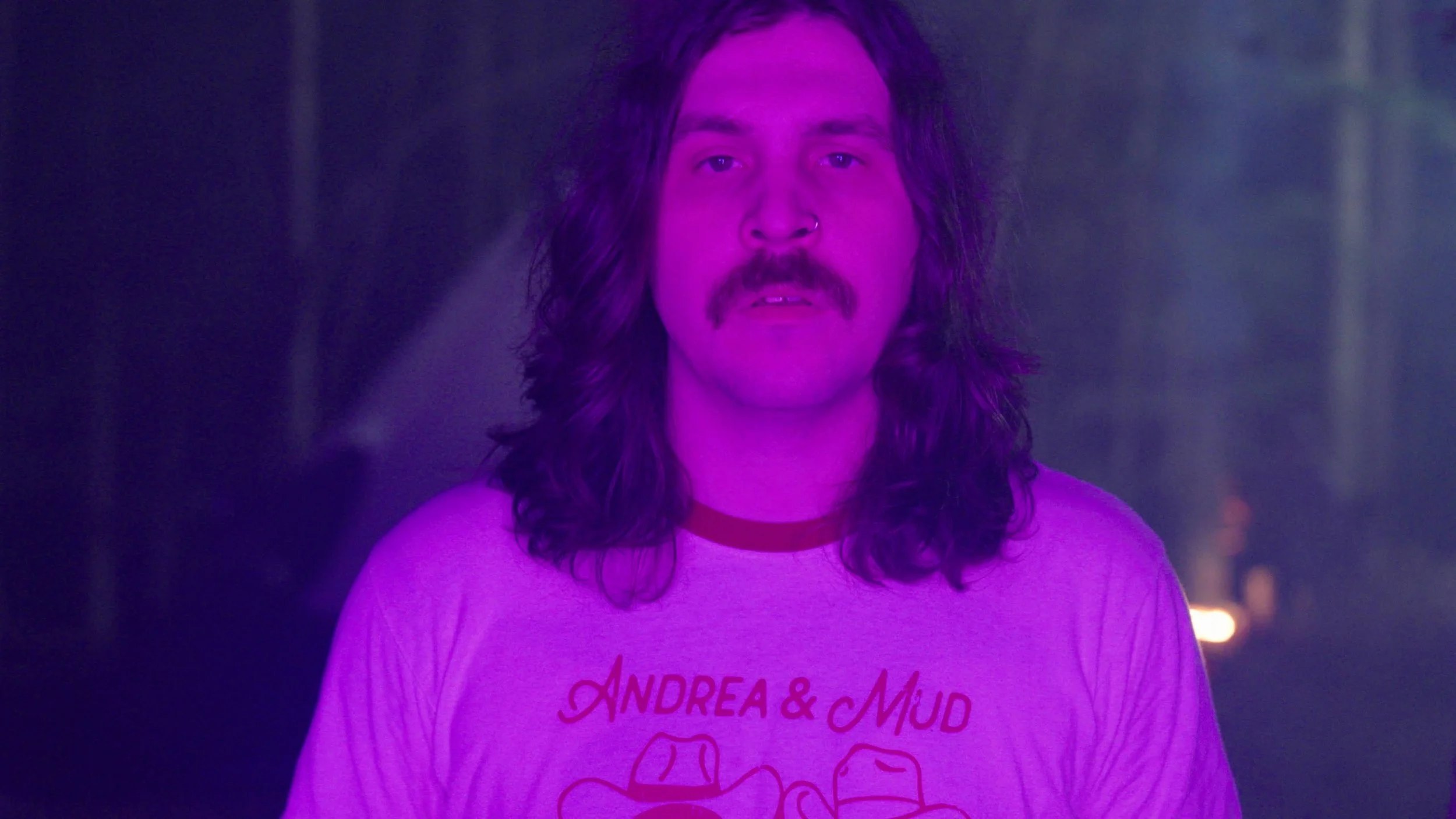 A man with long dark hair, a mustache, and a nose ring, wearing a T-shirt with the words 'Andrea & Mud' and a graphic of mules on it, standing in a dimly lit environment with purple lighting.