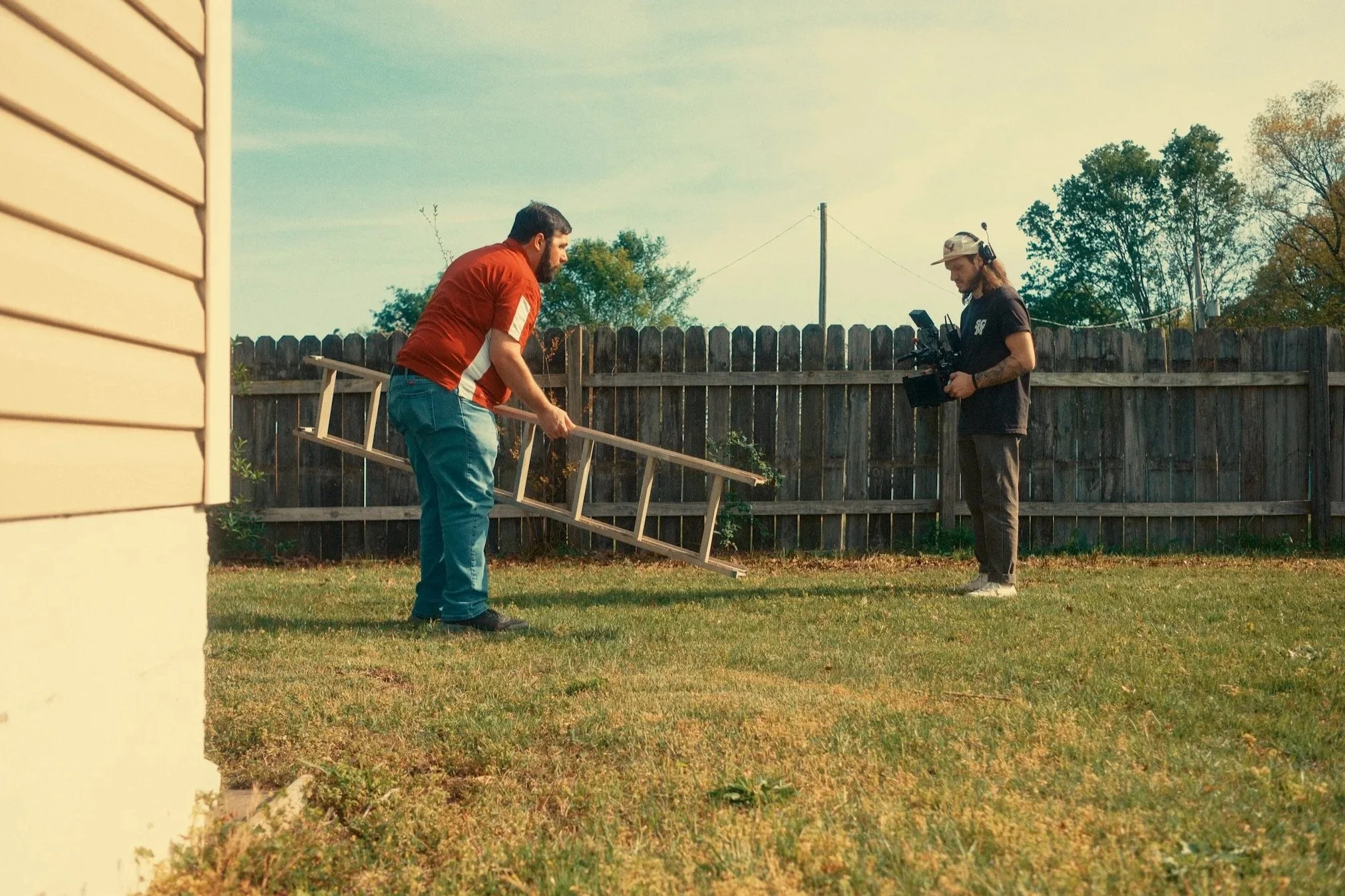 Two men setting up filming equipment in a backyard, with a wooden fence and trees in the background.