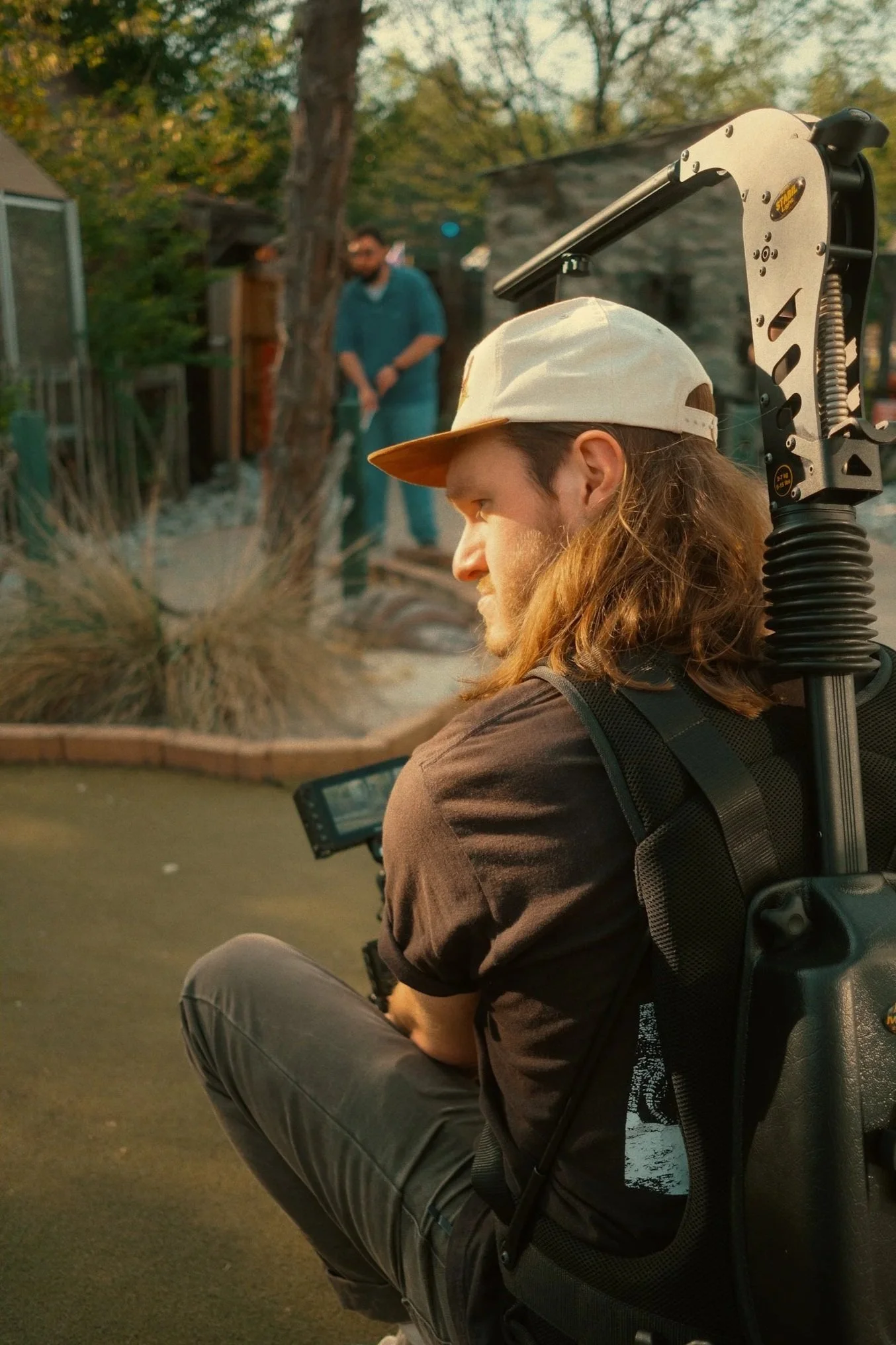 A man with long hair wearing a beige cap, sitting outdoors with a camera on a gimbal, watching a person in the background near a tree.