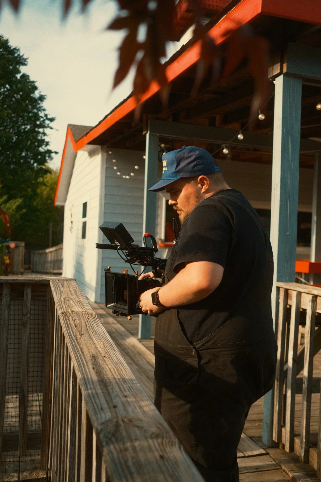 A man wearing a blue cap and black t-shirt standing on a wooden porch, holding a professional camera with a mounted monitor, with a small white building and string lights in the background.