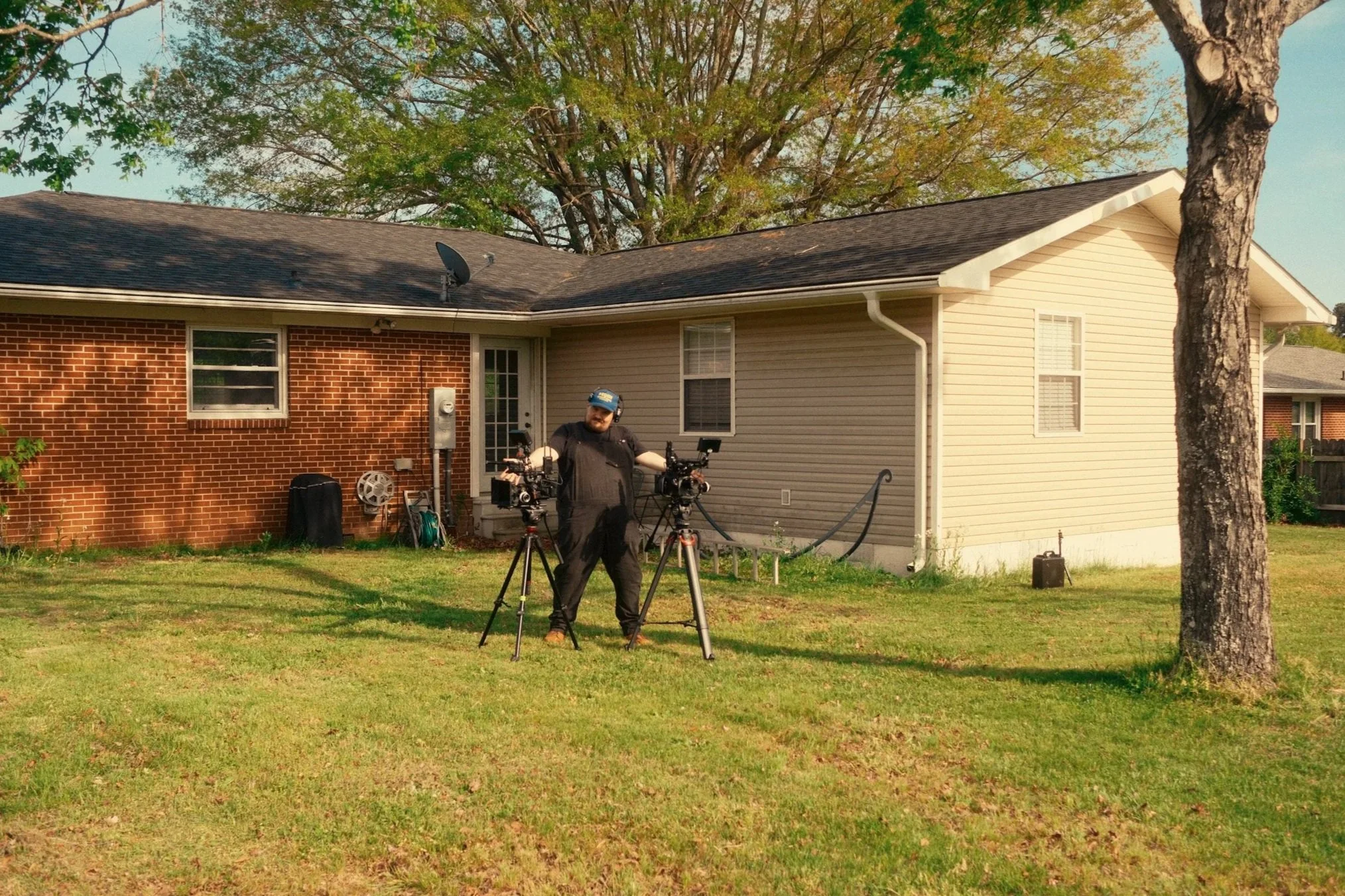 A man with a beard and dark clothing standing outside in a yard, operating dual professional film cameras mounted on tripods. Behind him is a house with a brick and beige siding exterior, trees, and a grassy lawn.