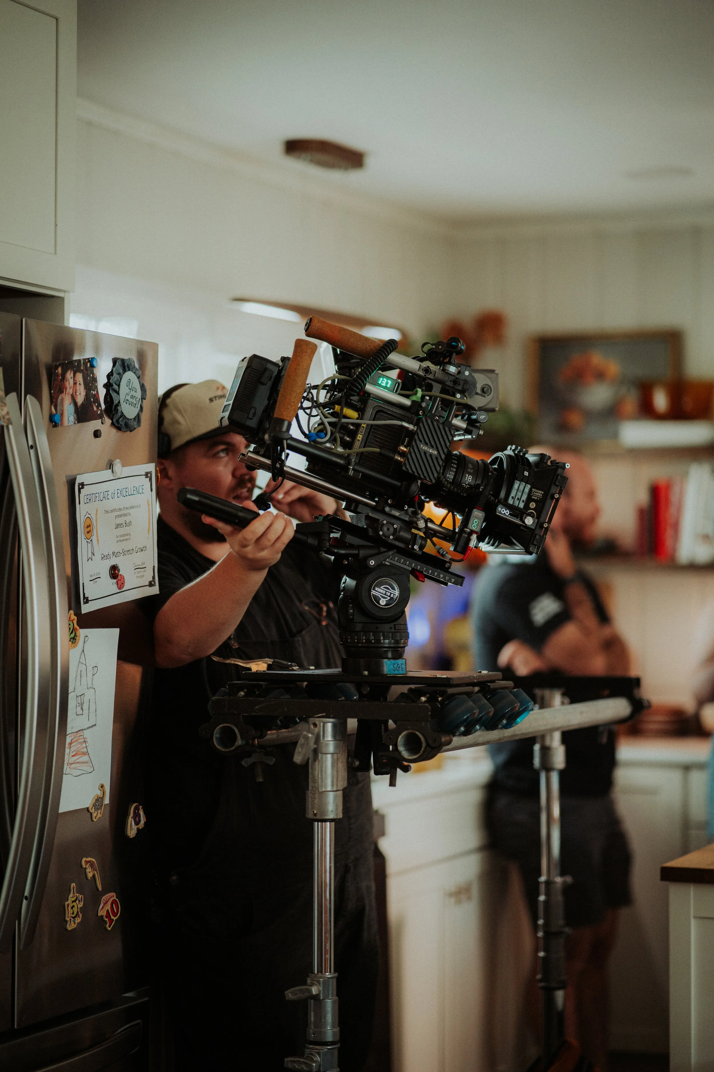 A camera operator films in a kitchen, with a woman in the background near a countertop.