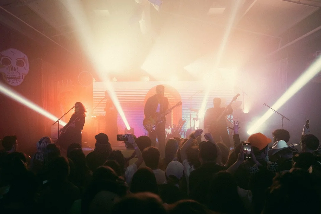 A live concert with a band performing on stage, colorful lighting, and a crowd of fans watching and taking pictures.