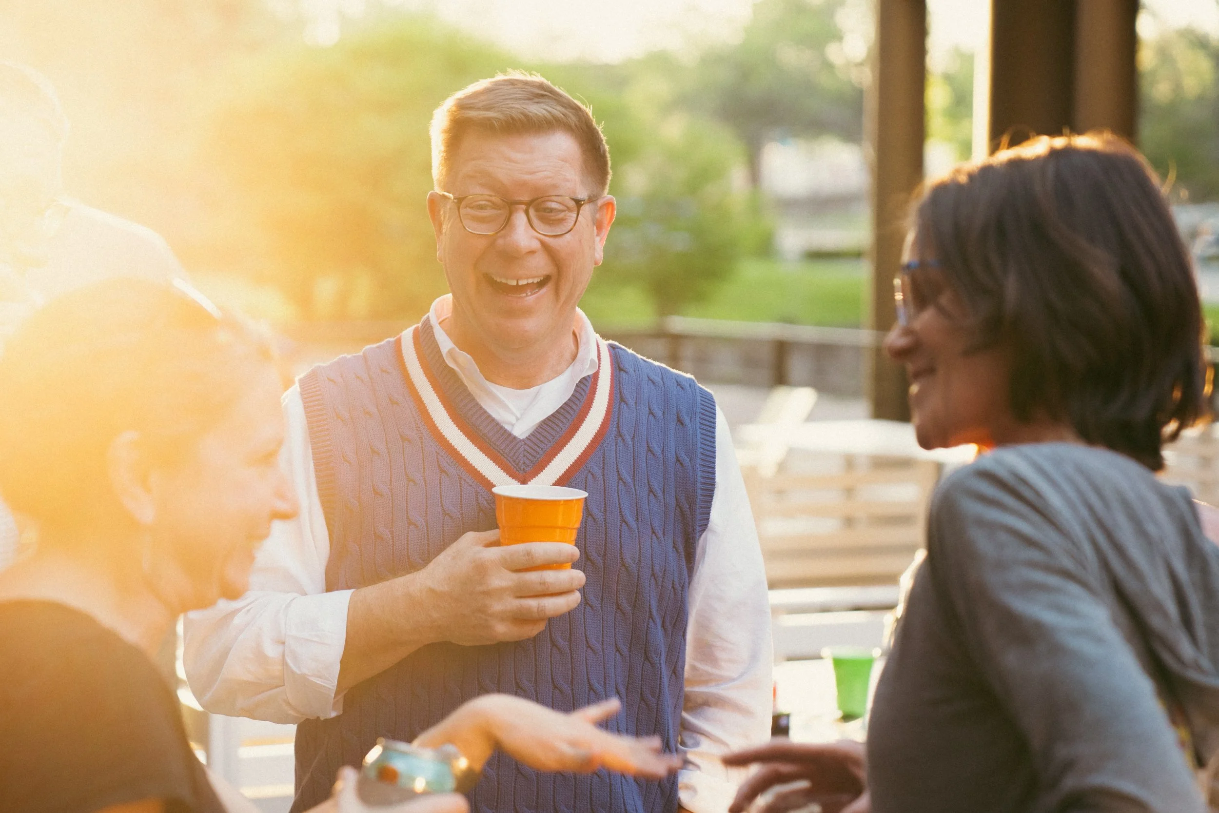 Three people happily talk outdoors during sunset, with trees and a porch in the background. The man in the center is wearing glasses and a blue sweater vest, holding a yellow cup, and smiling. Two women are on either side, engaging in conversation.