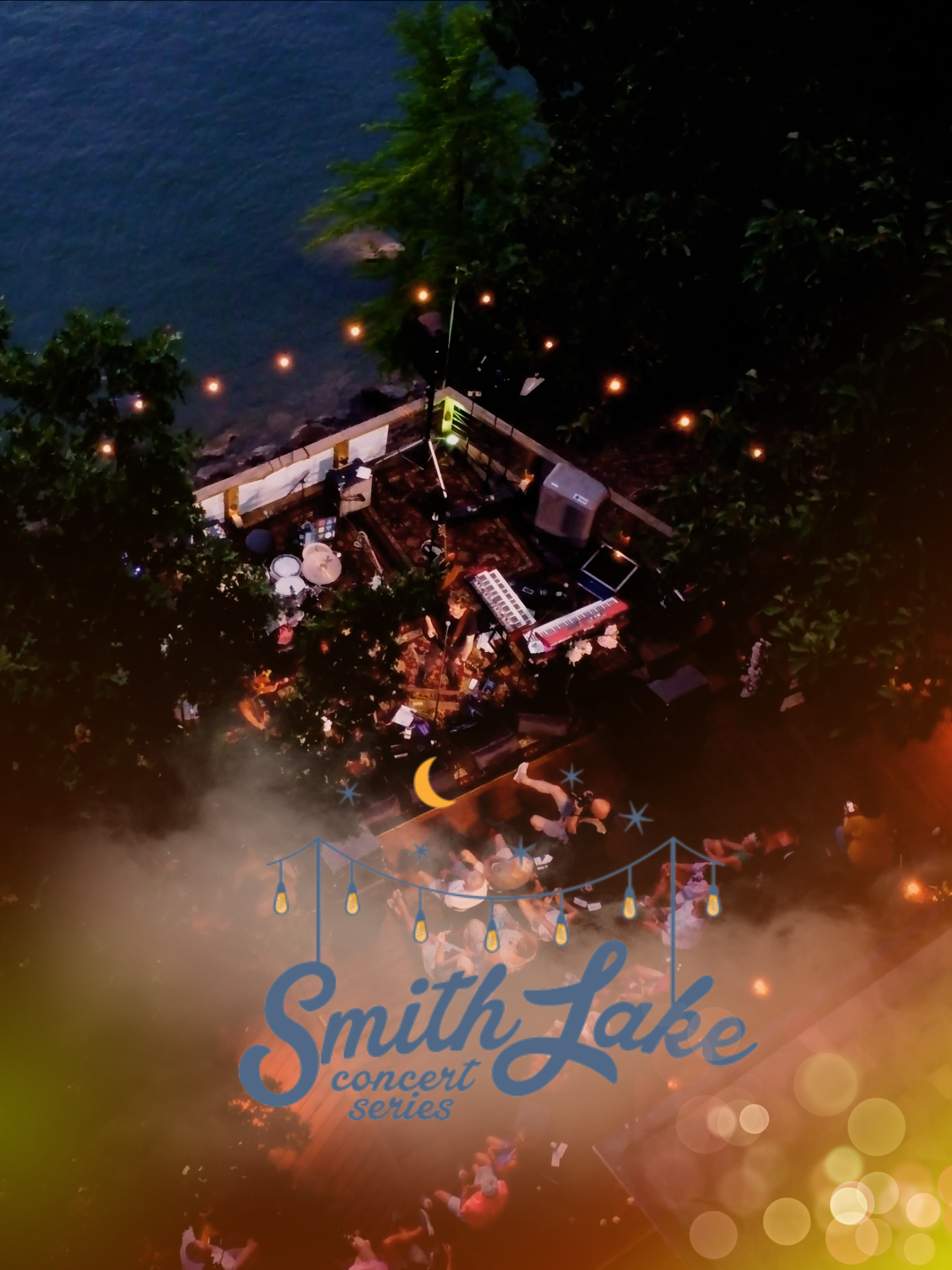 Bird's eye view of Smith Lake concert series at dusk, showing outdoor stage with musicians, audience seated around, string lights, and lake in background.