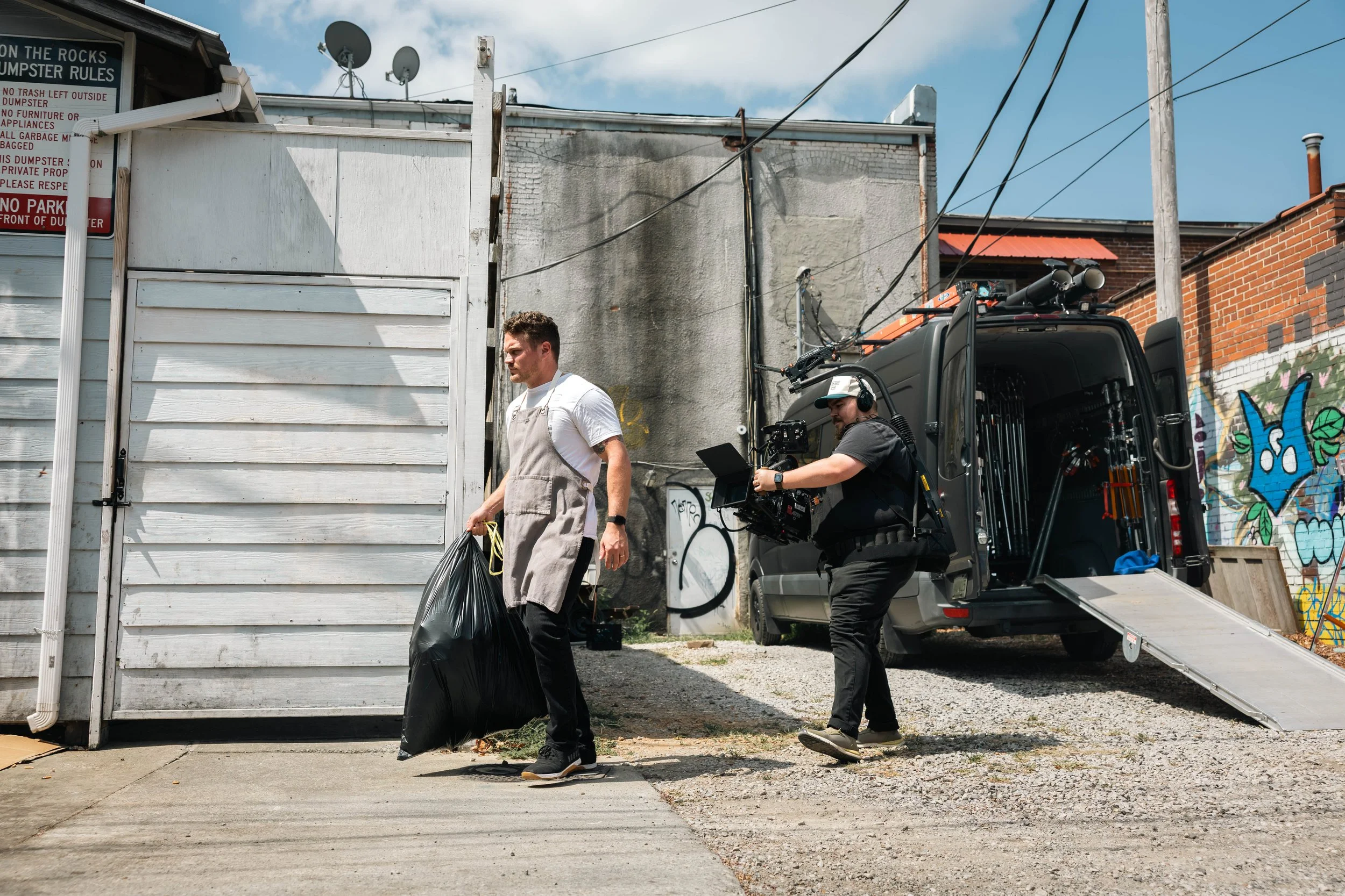 A man in an apron carries a black garbage bag outside a white building, with a film crew filming nearby. The crew member operates a camera on a rig, wearing headphones, and a black van with equipment is parked in the background, with graffiti on a br