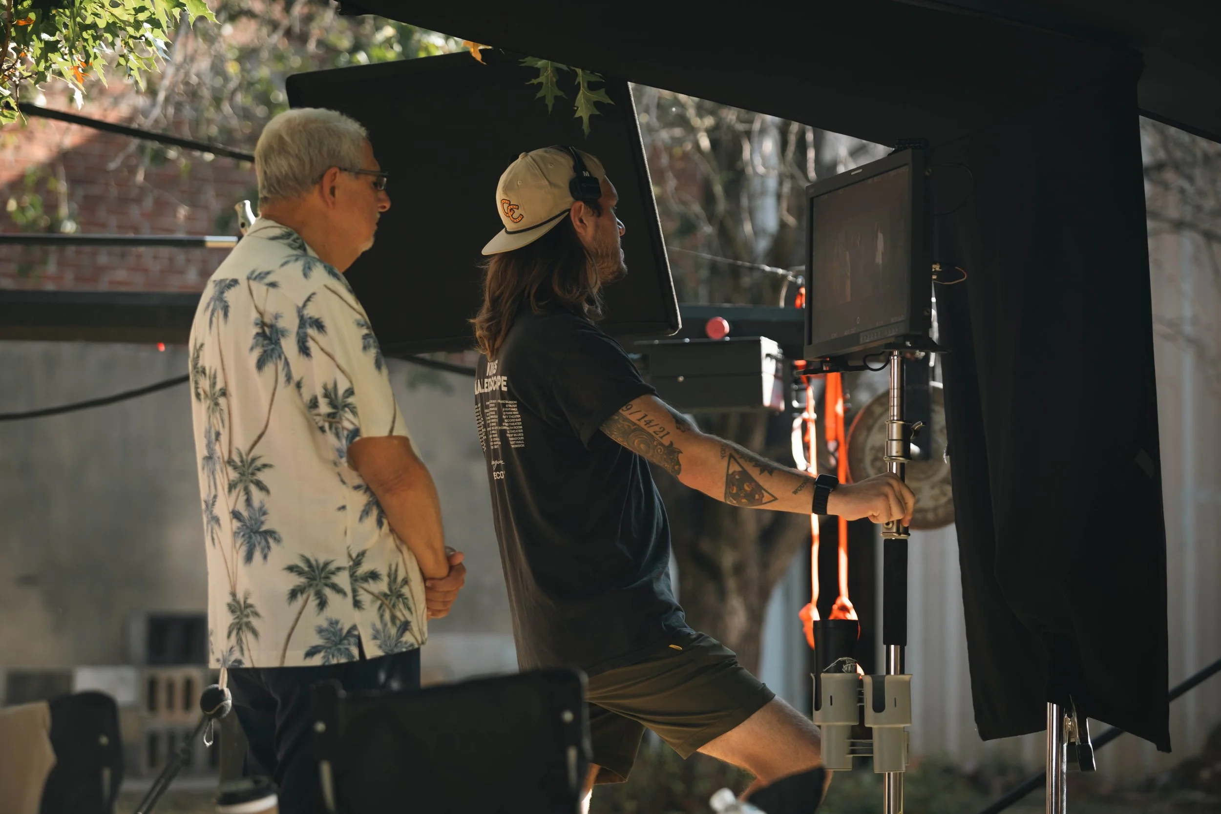 Two men working on a film shoot, one adjusting equipment on a stand and the other observing, outdoors with trees and a building in the background.
