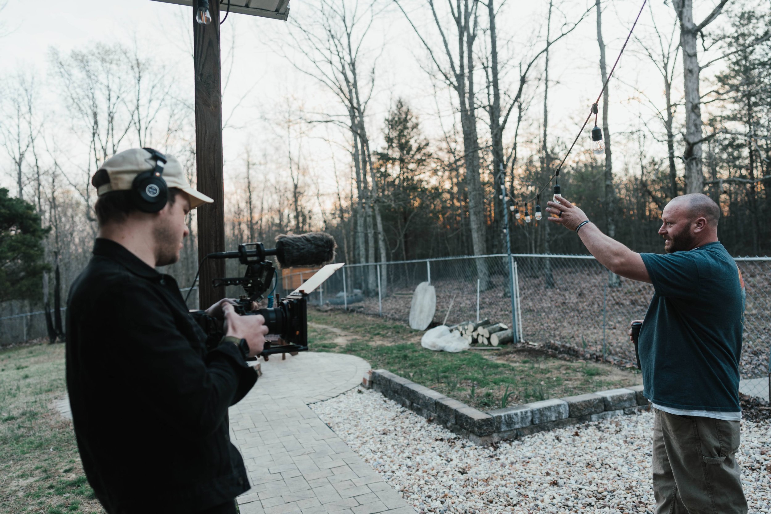 Two men filming a video outside during sunset, one holding a camera with a microphone, the other hanging string lights.