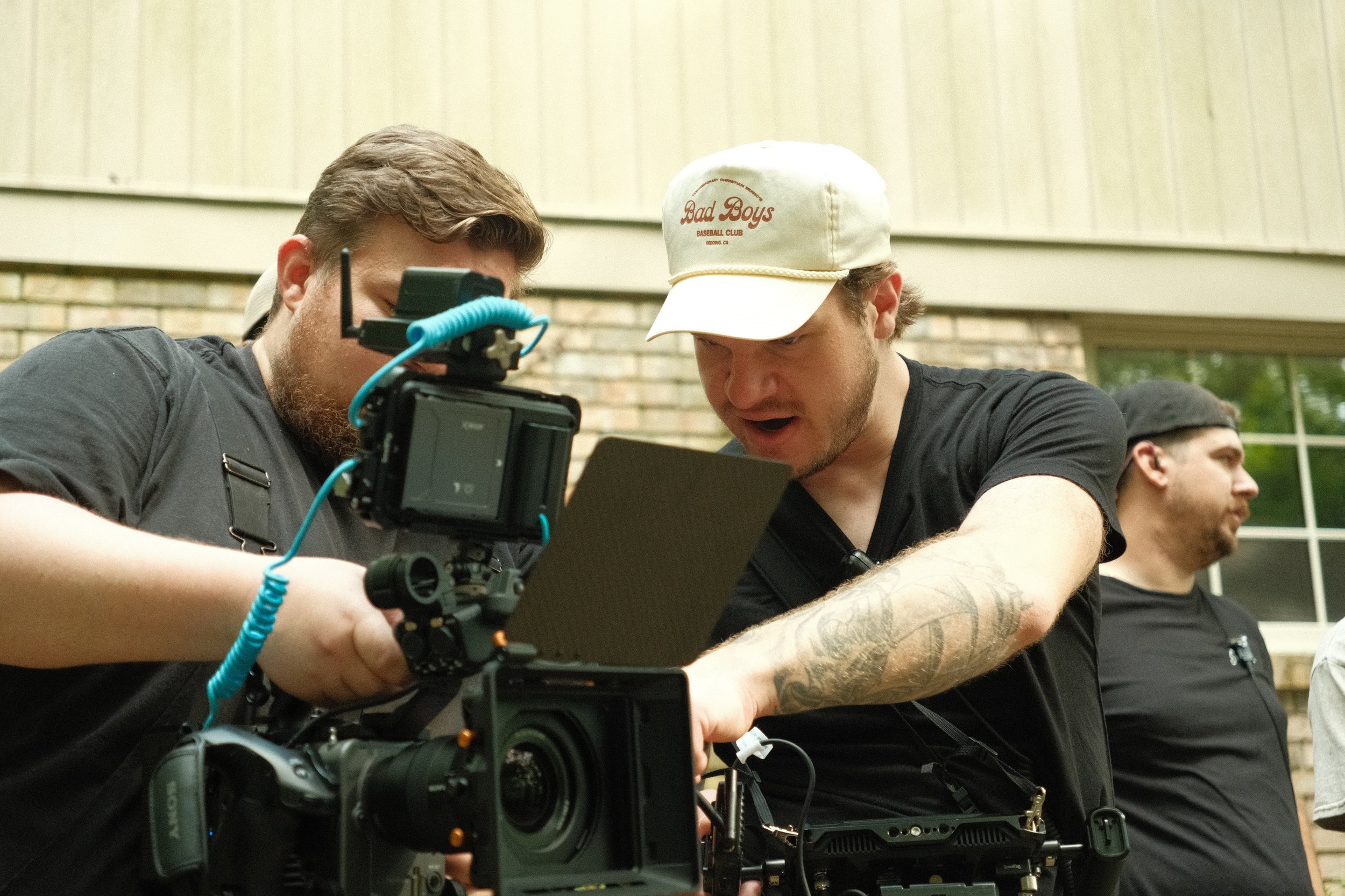 Two men working on a professional camera setup, with one man showing surprise or excitement, wearing a white cap that says 'Bad Boys Baseball Club,' and the other focused on adjusting equipment. A third man is standing in the background.