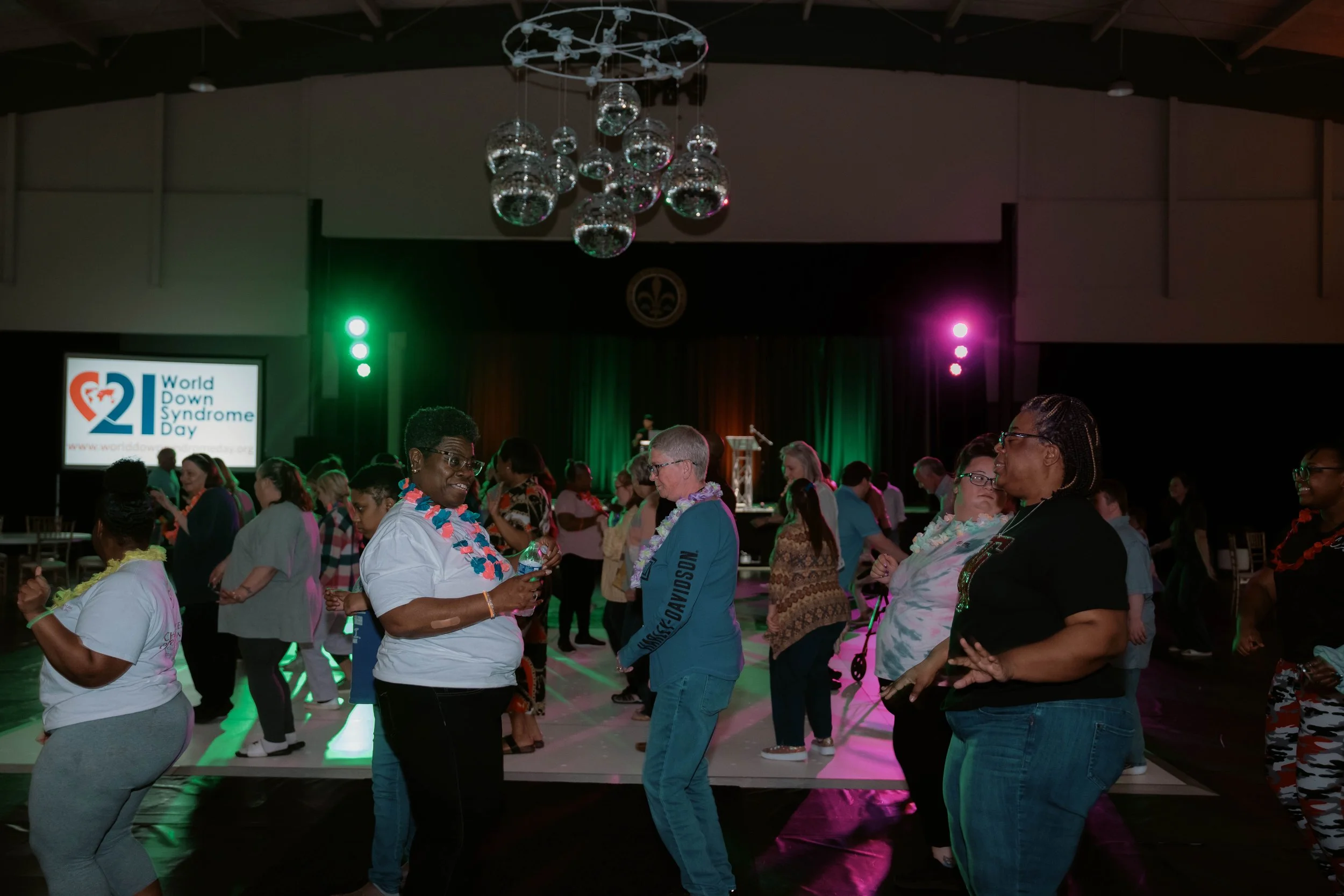 People dancing at an event for World Down Syndrome Day with a large screen displaying the event logo and a stage in the background.
