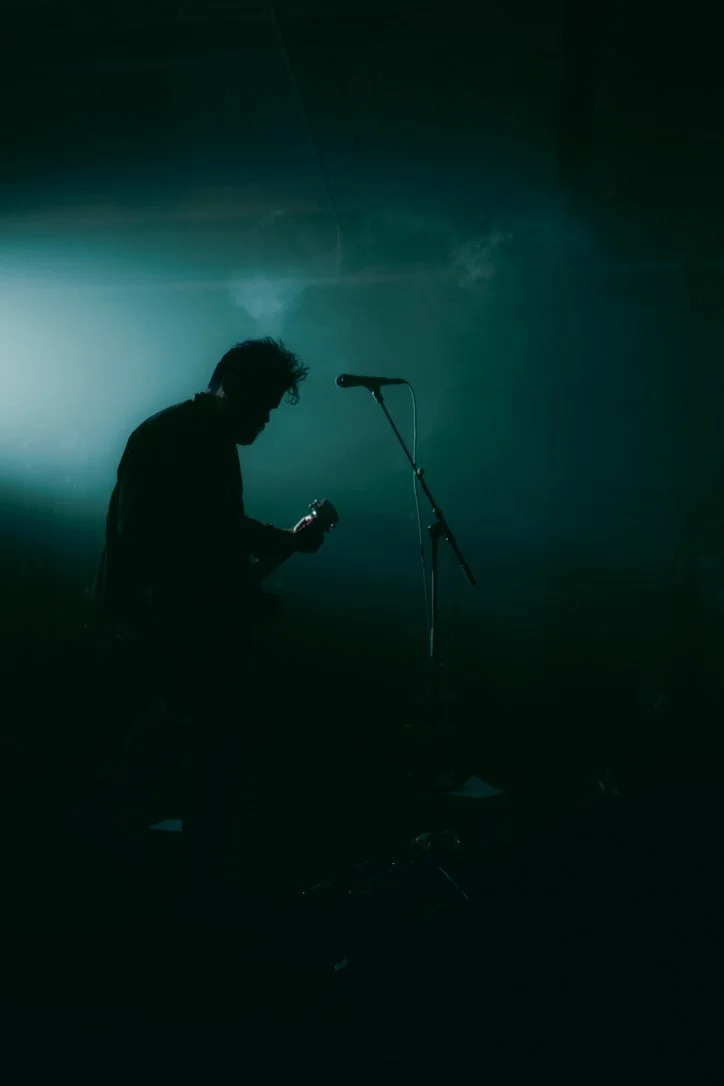 Silhouette of a musician holding a guitar in a dark stage with a microphone stand in front, illuminated by blue-green stage lighting.