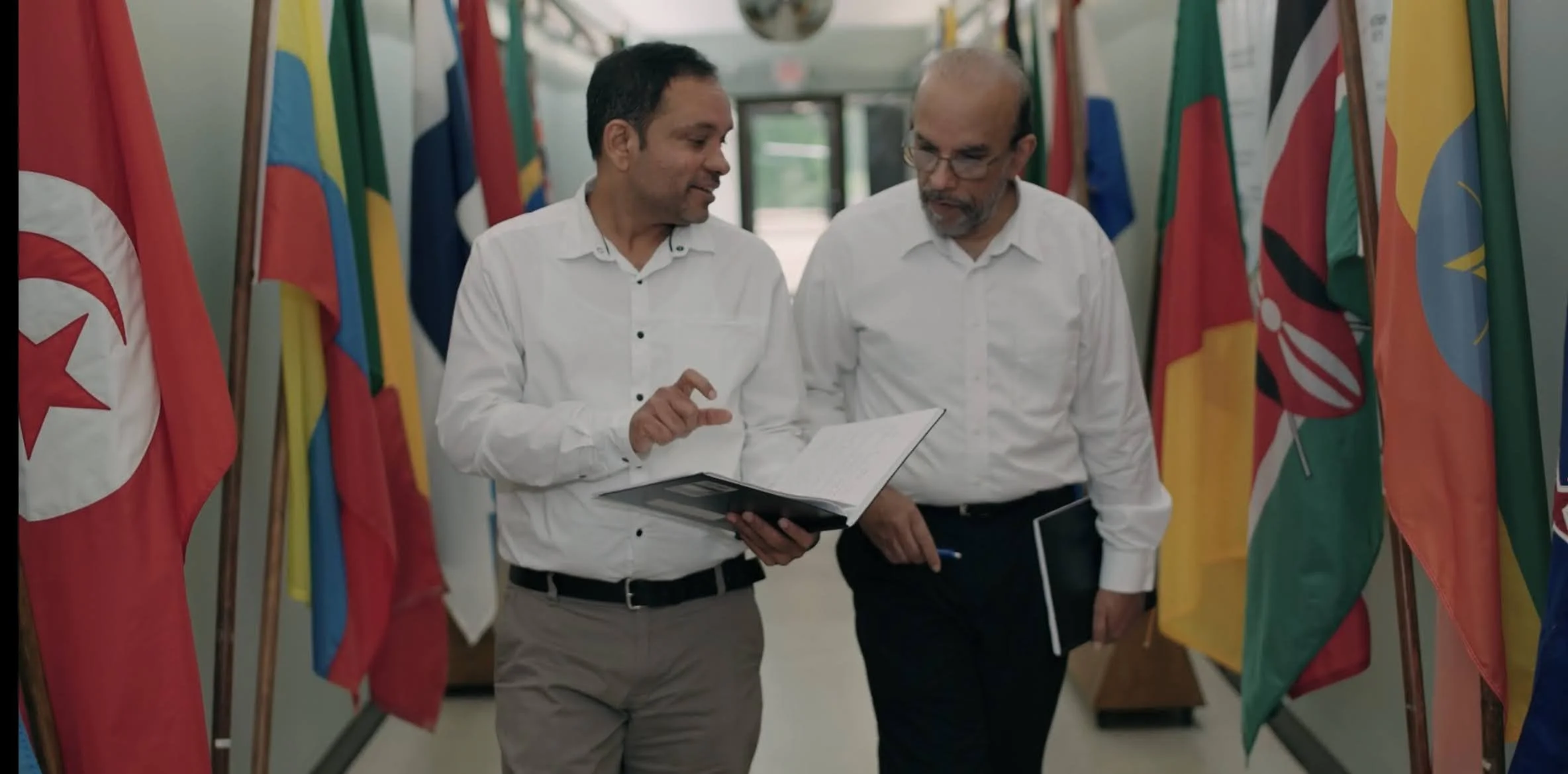 Two men in white shirts walking and talking down a hallway with various national flags displayed on each side.