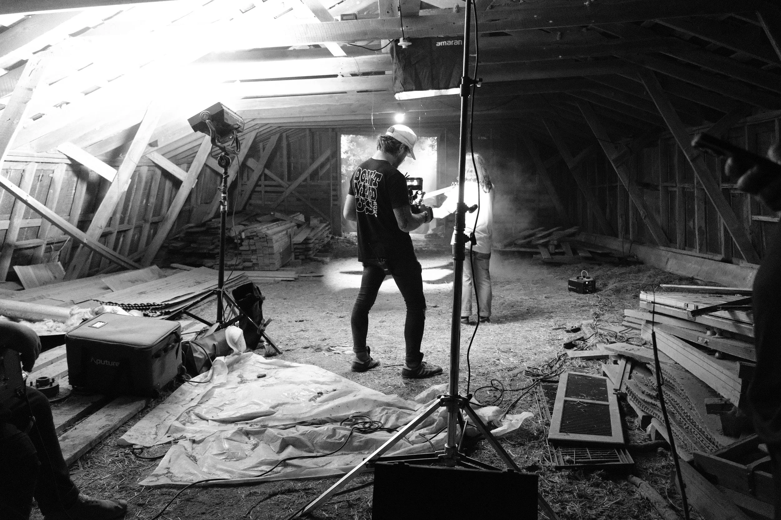 Inside a barn with wooden walls, a crew is filming a scene with professional lighting and cameras, showing crew members and equipment.