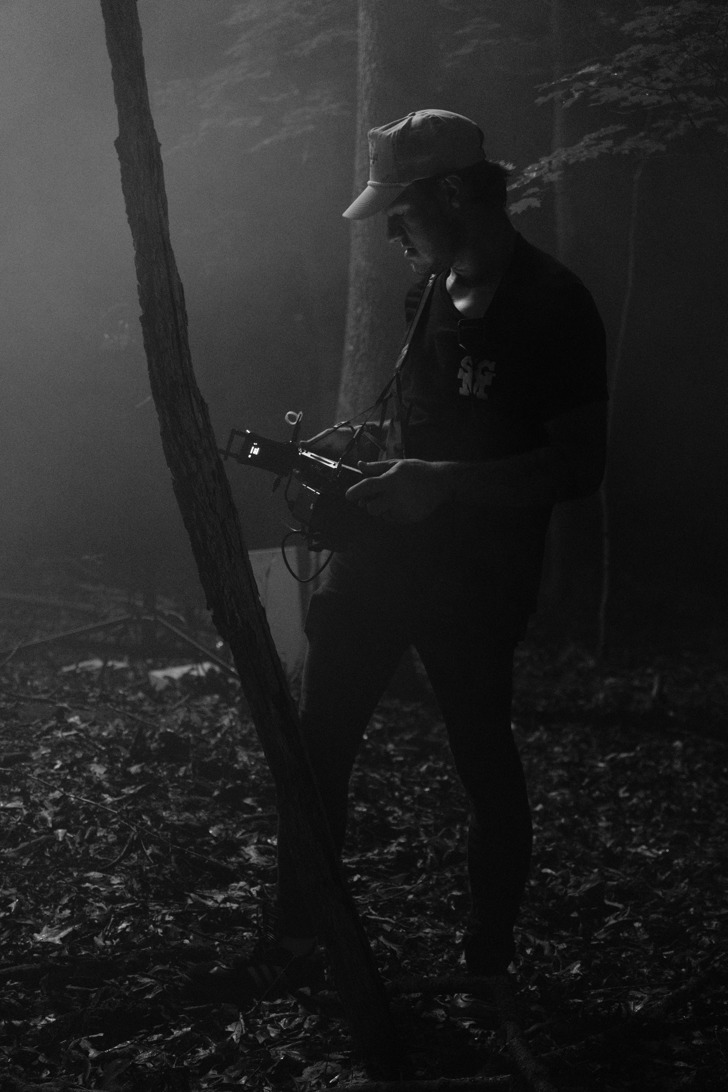 A person wearing a baseball cap stands in a forest, holding a camera or some electronic device, surrounded by trees and fallen leaves, with a smoky or foggy atmosphere.