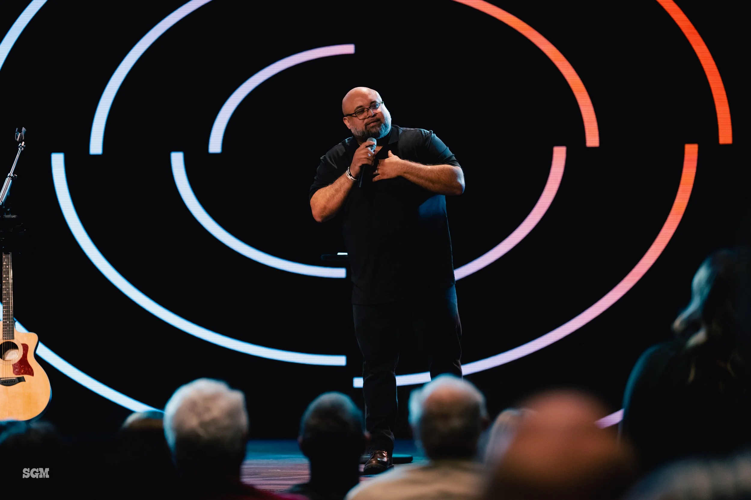 A man with a beard and glasses stands on stage holding a microphone, gesturing to his chest, with an audience in front and a guitar on a stand to his left. The background features concentric circular light patterns in blue, purple, and red on a black
