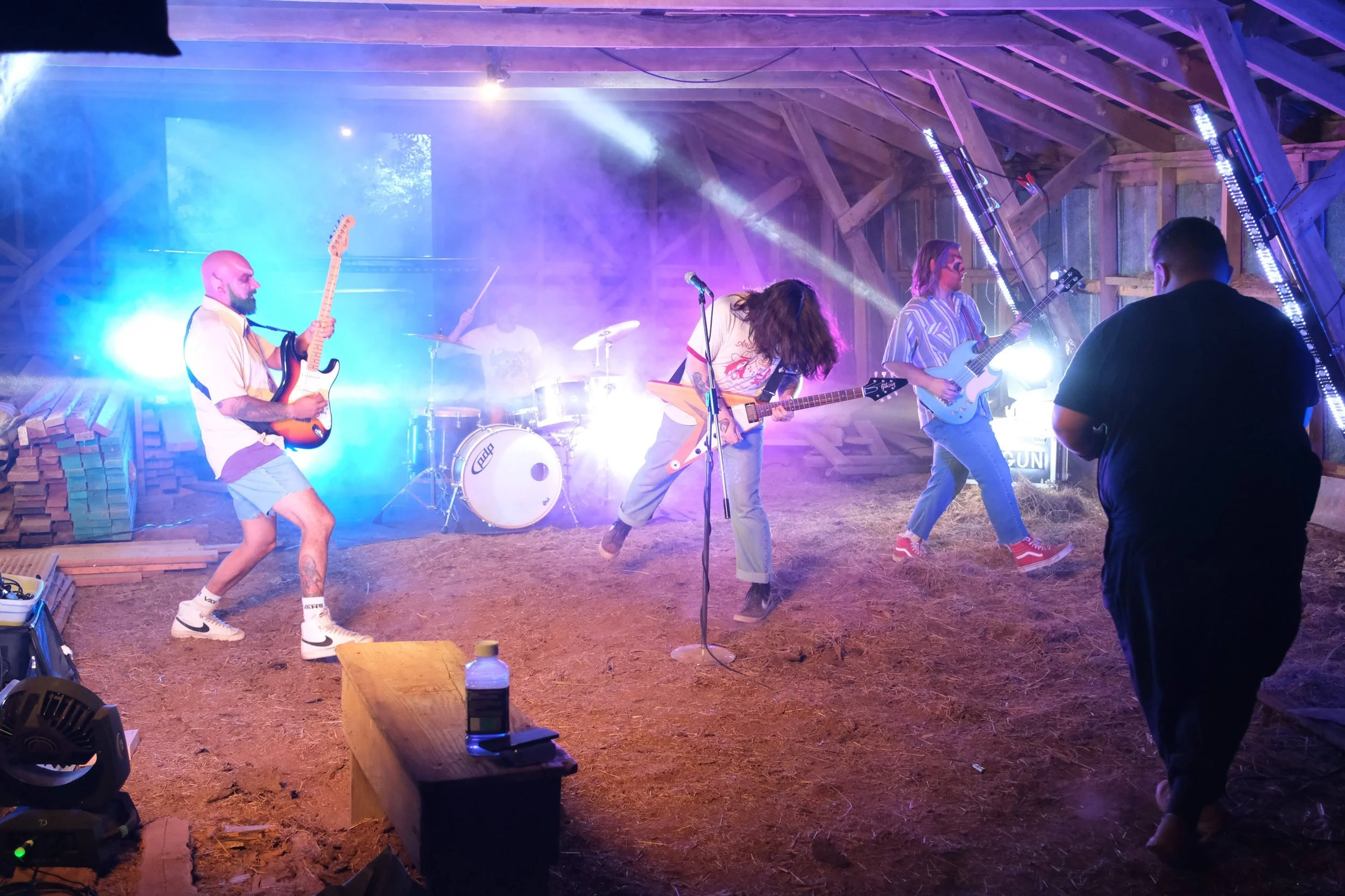 A band of five members performing passionately on stage inside a barn or rustic wooden structure with dynamic purple and blue stage lighting and fog effects.