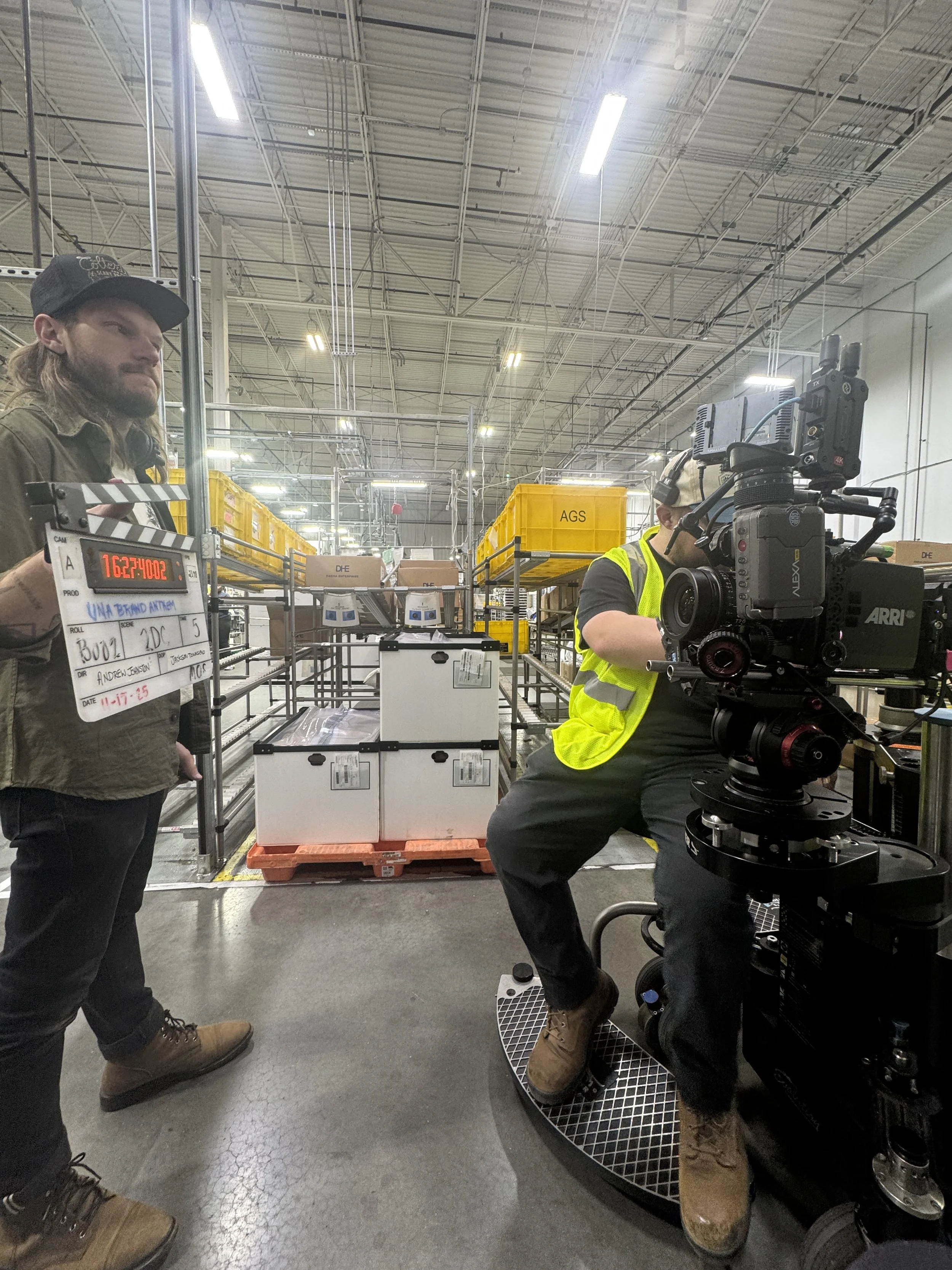 Two crew members working in a warehouse, one operating a camera mounted on a dolly while the other stands nearby. Warehouse shelves with yellow bins and boxed items are visible in the background.