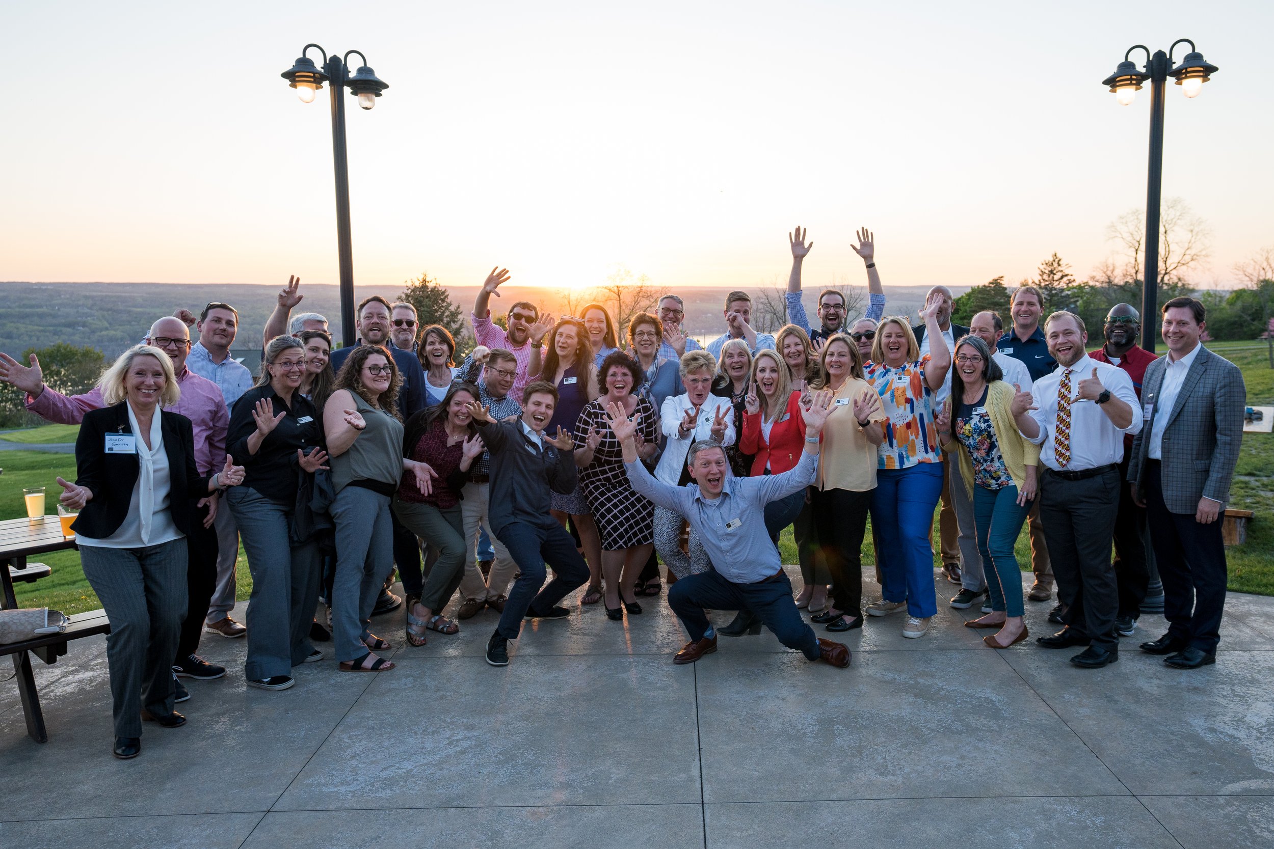 A large group of people gathered outdoors at sunset, smiling and waving at the camera, with scenic hills and two street lamps in the background.