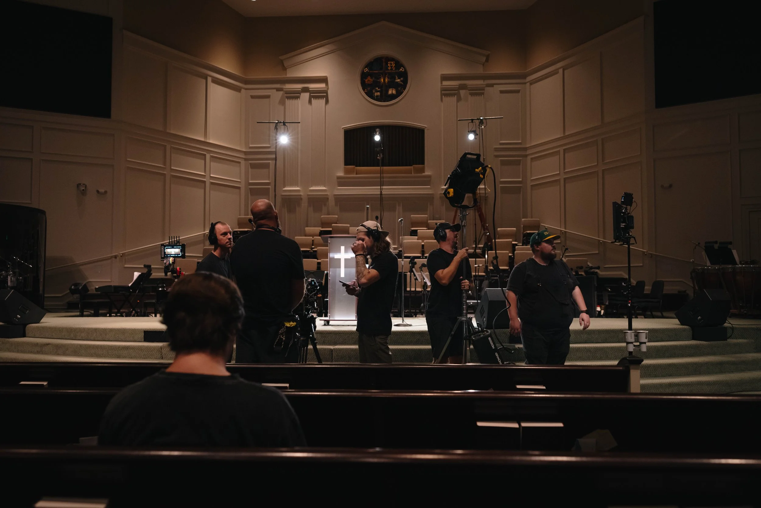 Film crew setting up equipment in a church interior with a cross on a lectern at the front.