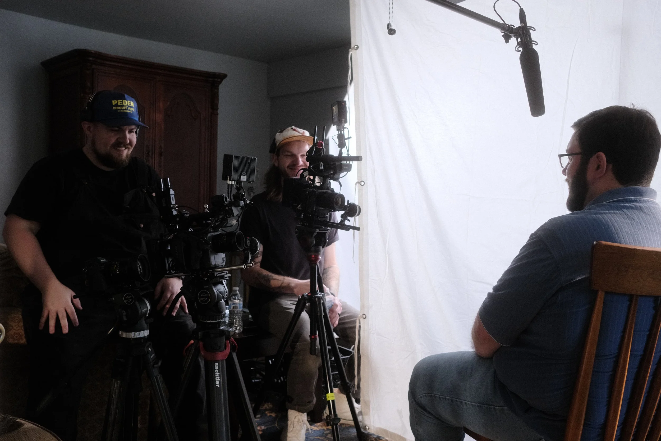 Two camera operators filming a man with glasses and a beard sitting on a wooden chair against a white backdrop in a room with a wooden cabinet.