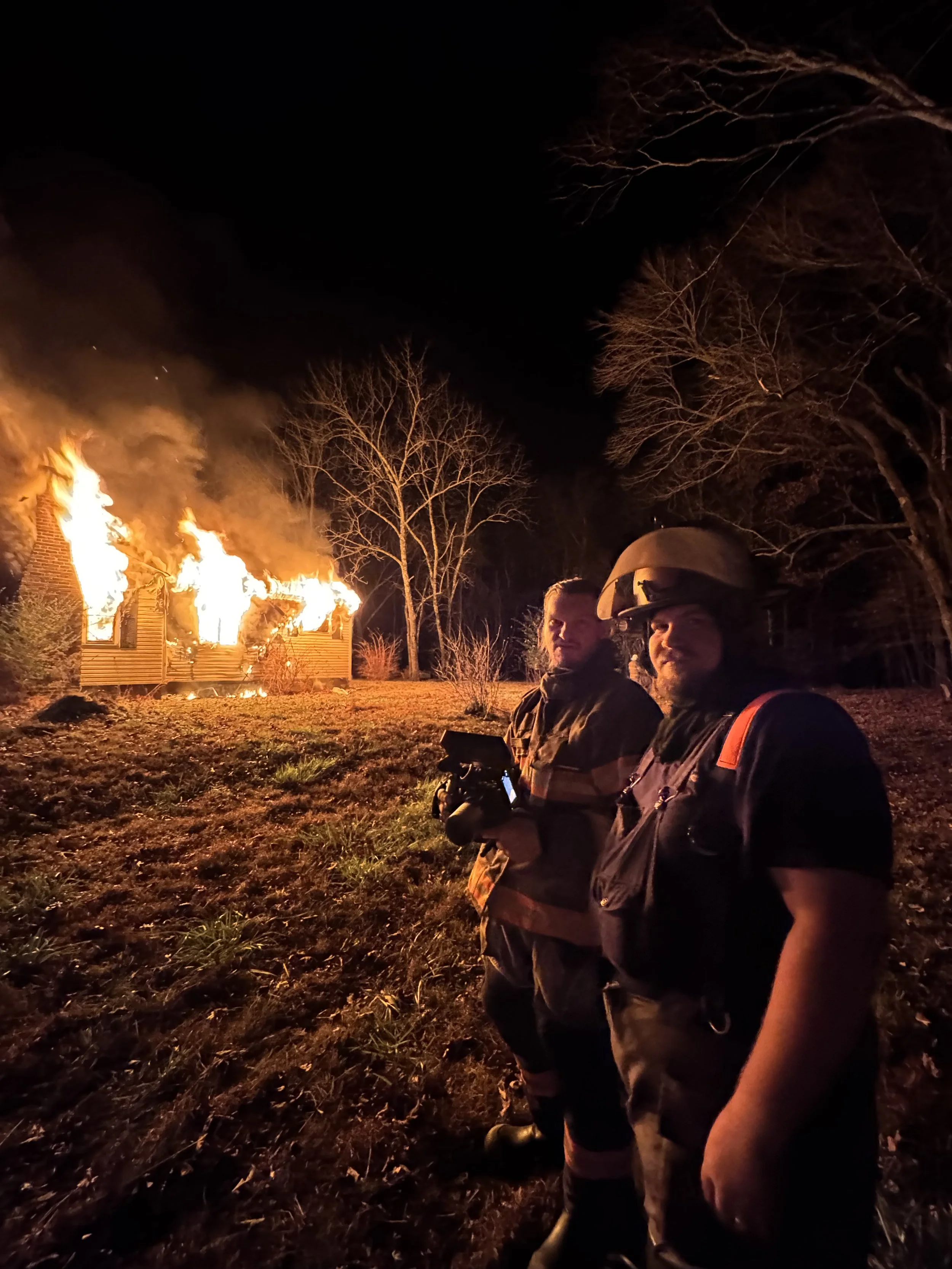Two firefighters stand outside at night with a house engulfed in flames behind them.
