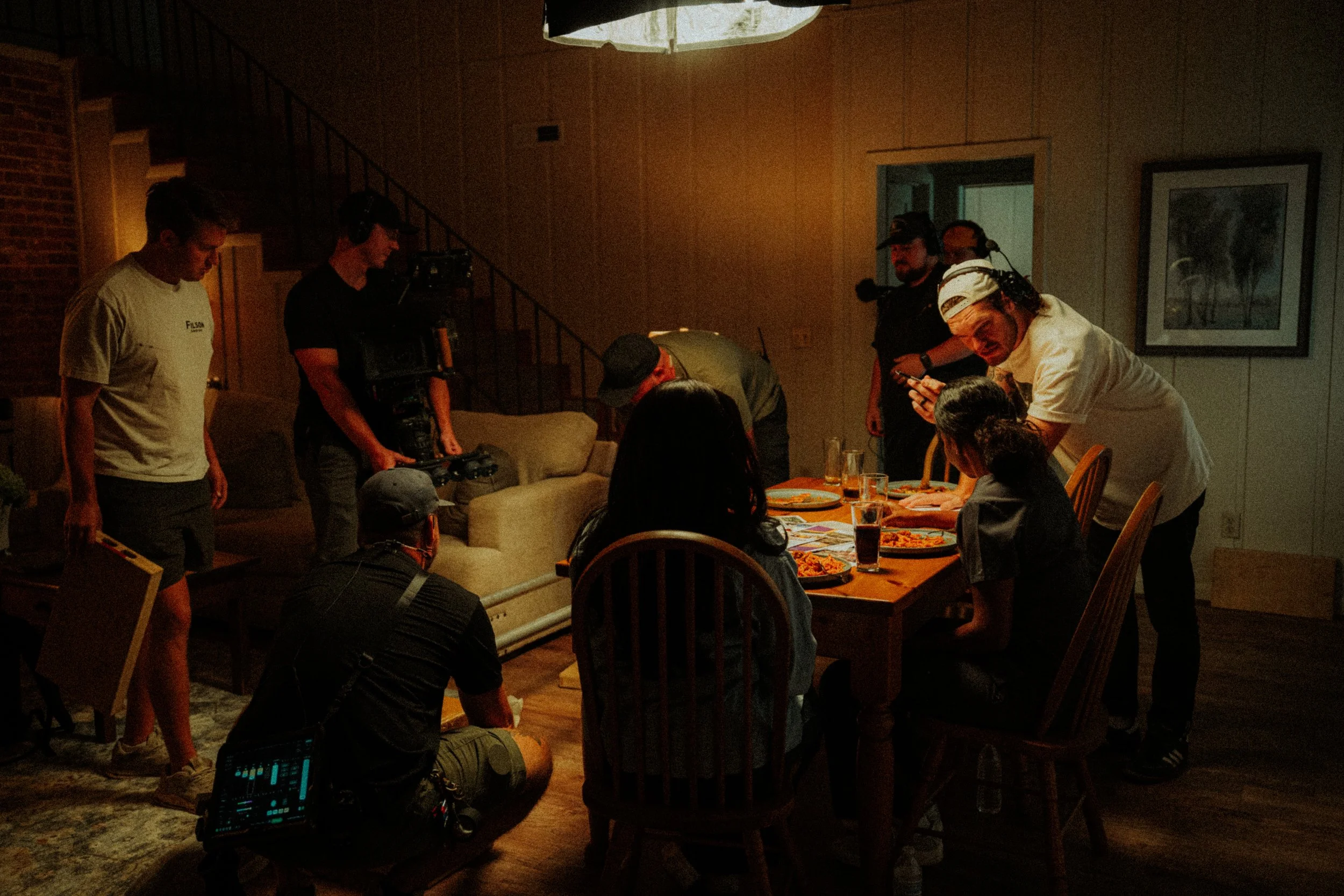 A film crew working on a scene in a dimly lit room, filming a woman at a dining table with pizza, with crew members operating cameras and equipment.