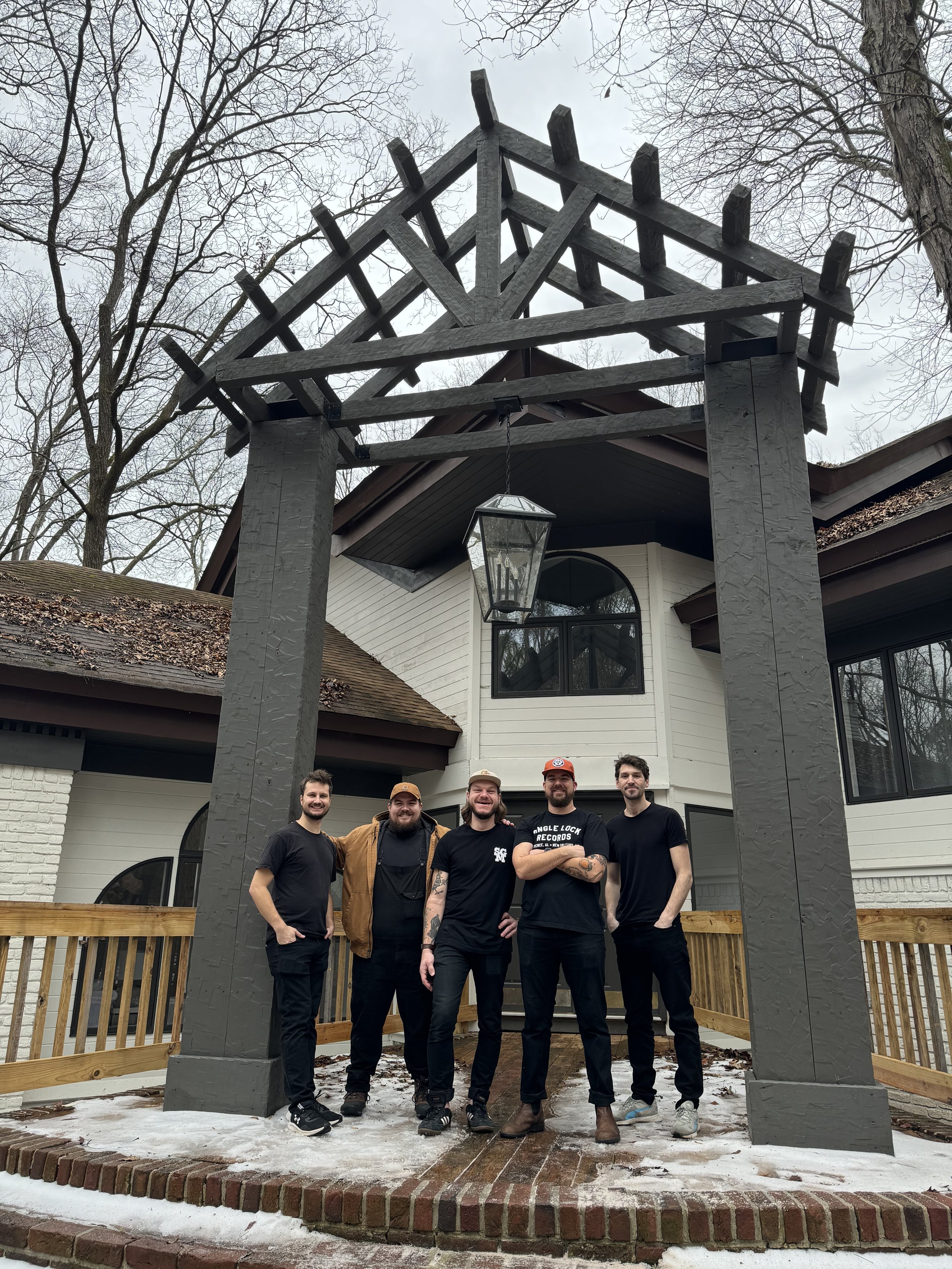 Group of five men standing on a brick and wooden porch in front of a large white house with black accents and a timber frame entrance, during winter with snow on the ground and leafless trees in the background.