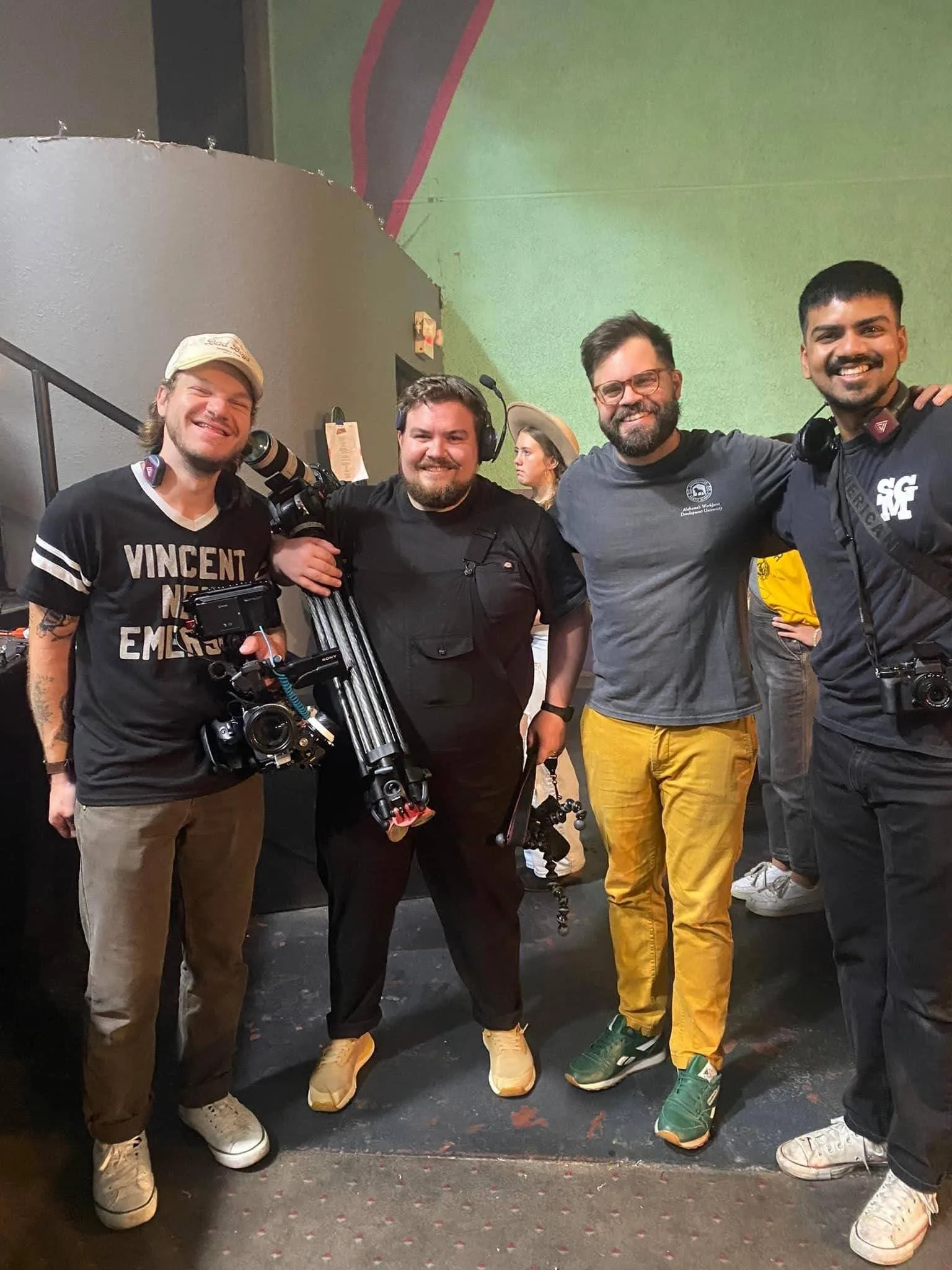 Four men standing together in a film set, smiling at the camera, with cameras and gear around them.