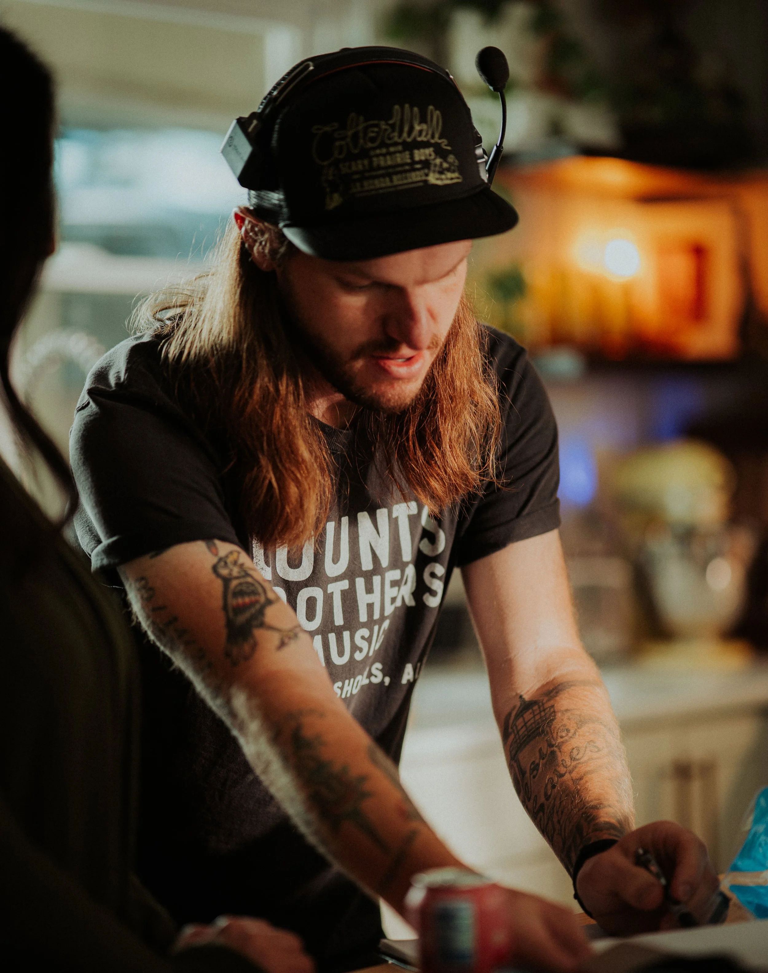 A man with long hair, tattoos, wearing a black cap with a headset, writing on a piece of paper in a kitchen.