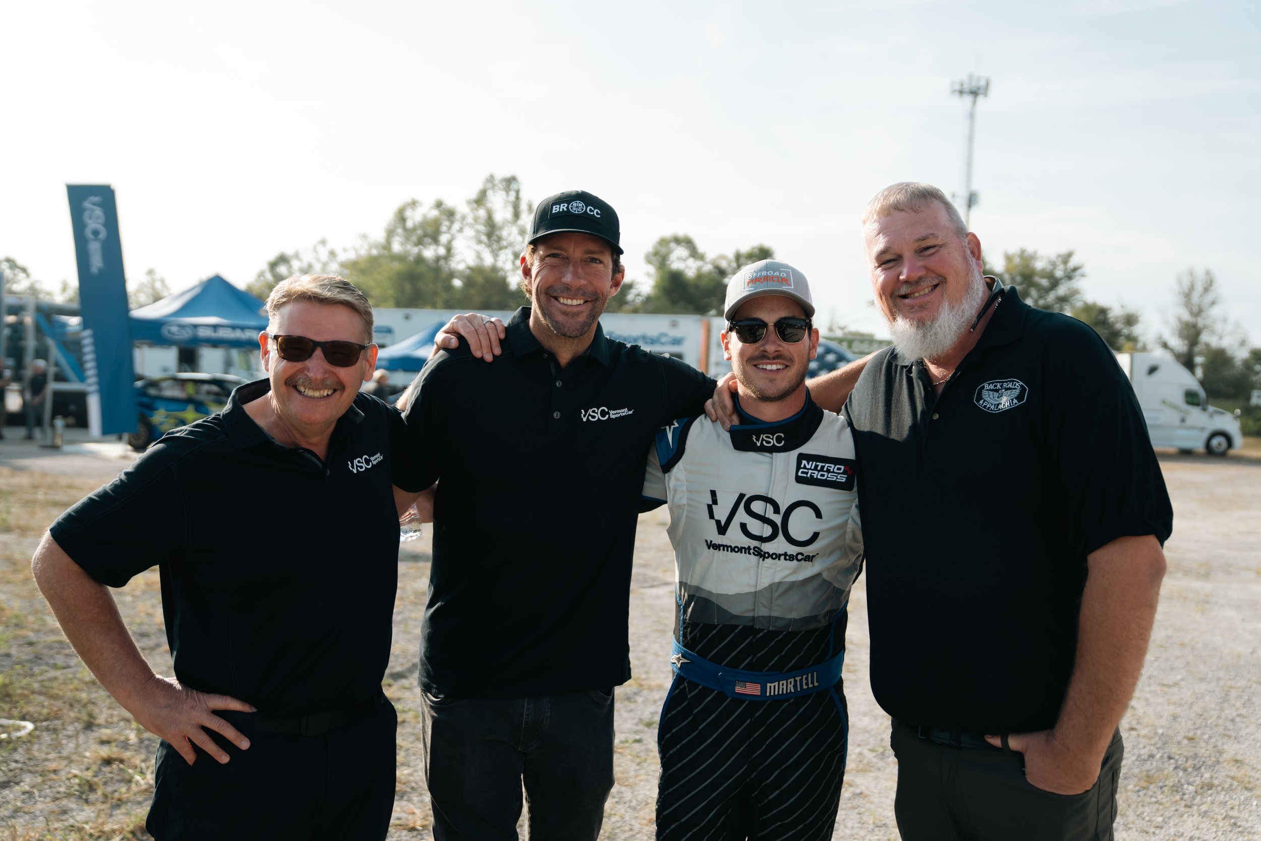 Four men smiling in racing gear and team shirts, standing outdoors with racing trailers and tents in the background.