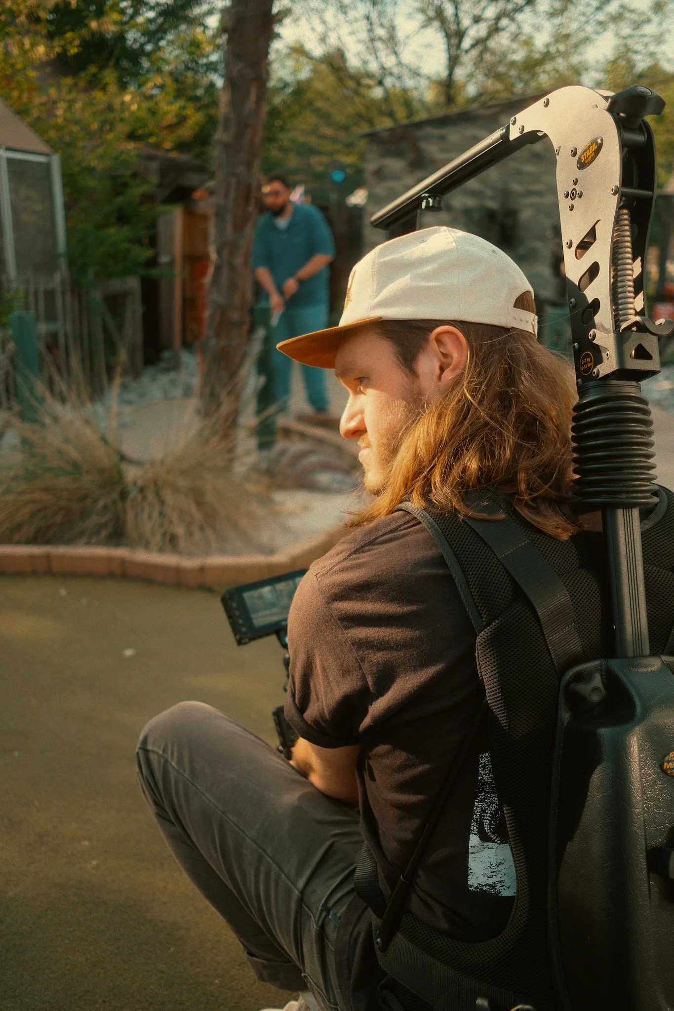 A young man with long hair, wearing a cap backward, sitting outdoors with a camera on a stabilizer on his back, filming a scene involving another person in the background near some trees and landscaping.