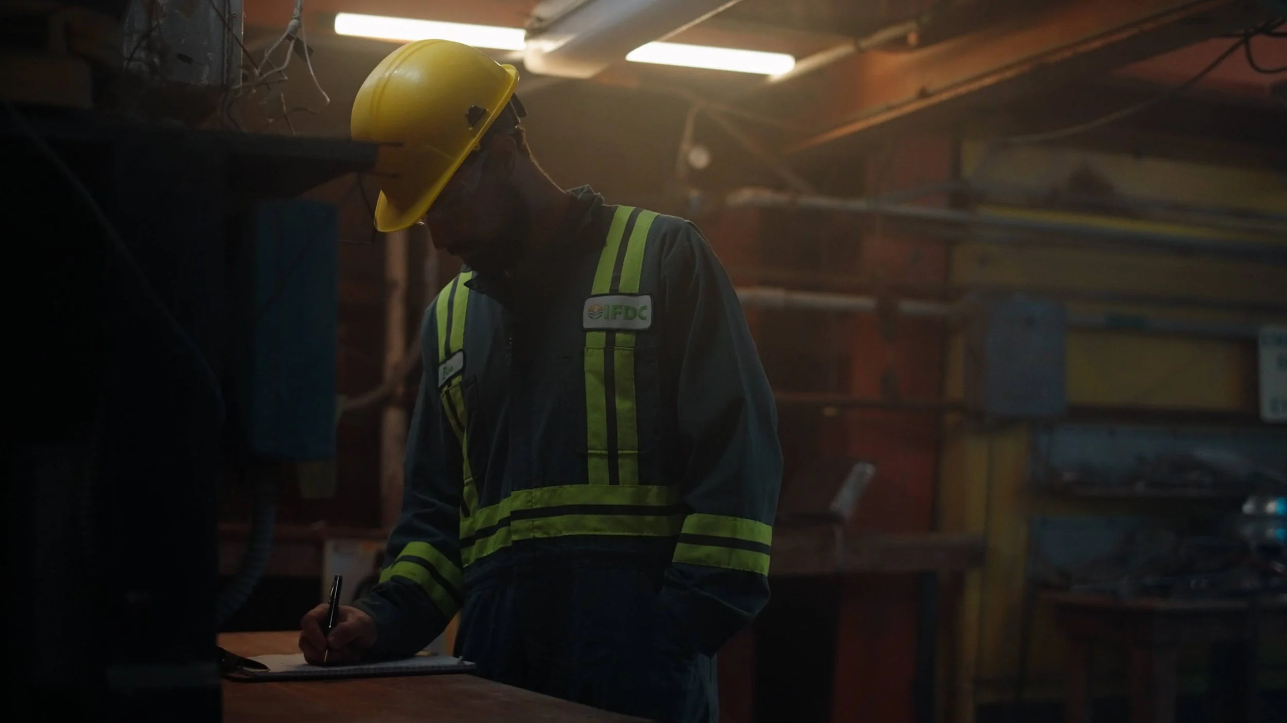 A construction worker wearing a yellow safety helmet and a dark jacket with reflective stripes is writing on a clipboard in a dimly lit industrial environment.