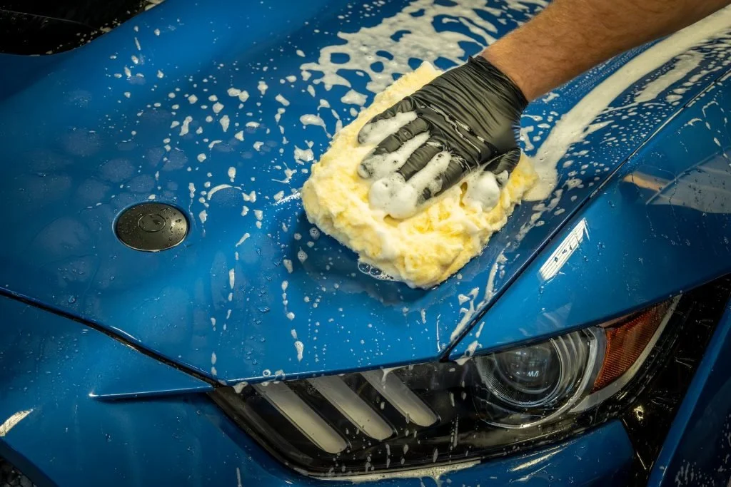 Person in a black glove washing a blue car with a yellow sponge and soap suds.