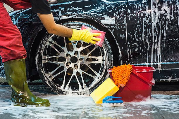 Person washing a black car's wheel with soap and sponge, wearing yellow gloves and green rain boots, with cleaning supplies like a red bucket, a yellow sponge, and a blue scrub brush nearby.