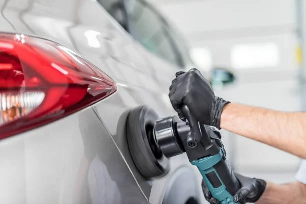 Person polishing the side of a gray car with a power buffer in a garage.