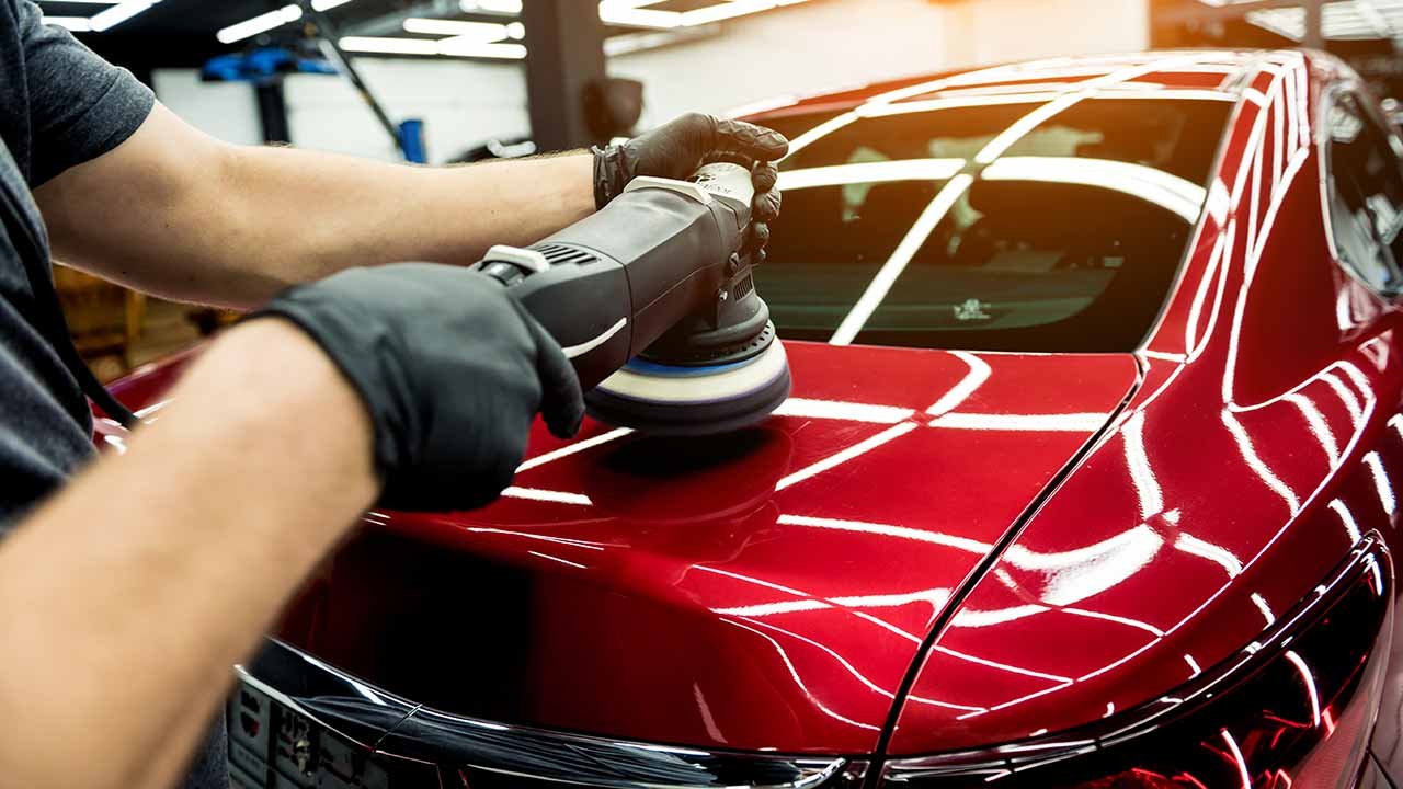 Person polishing a shiny red car with a power buffing machine in an indoor garage.
