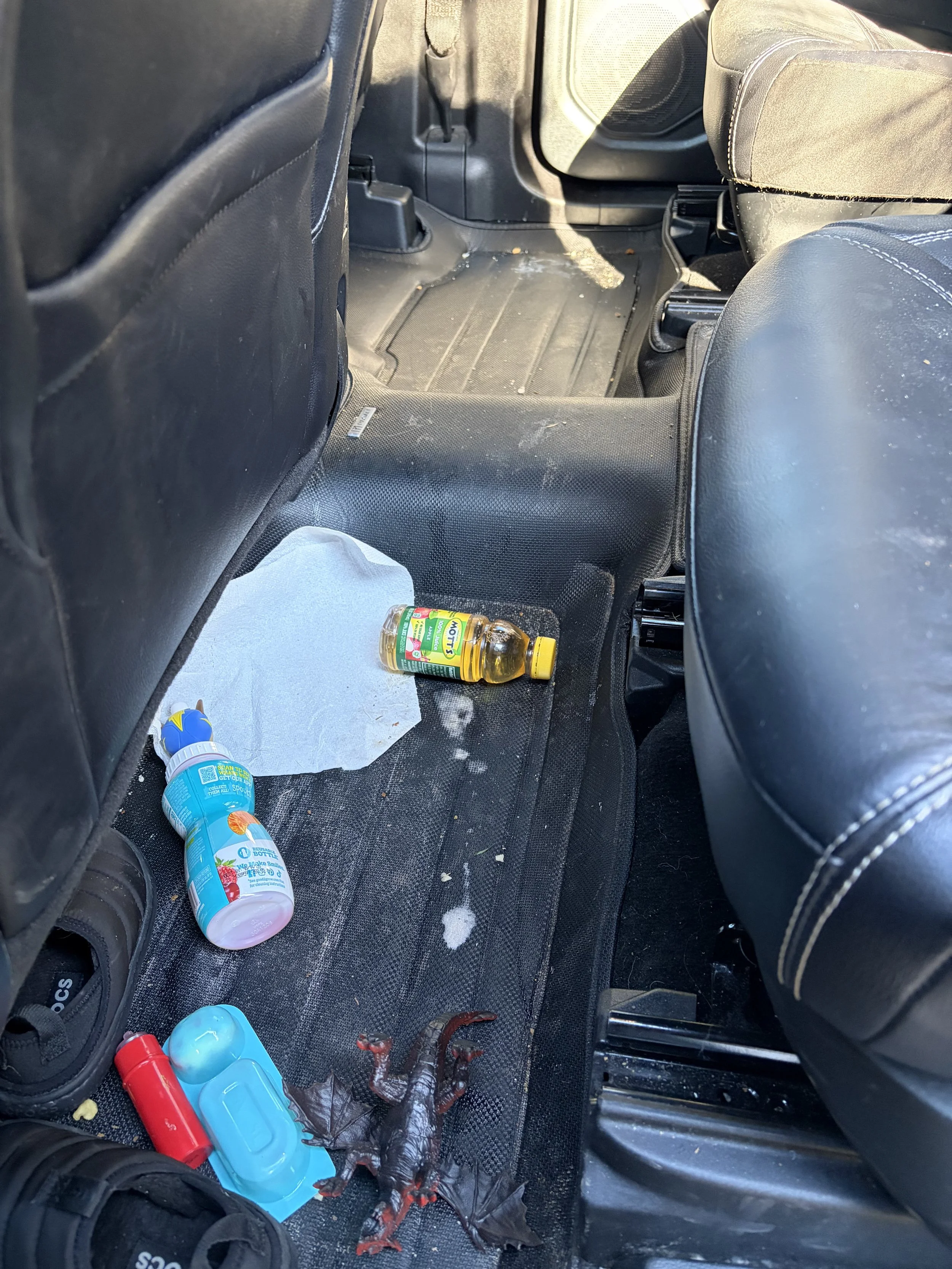 The back passenger area of a car with some toys, a drink bottle, a leaf, and a toy dinosaur on the dirty floor mat.