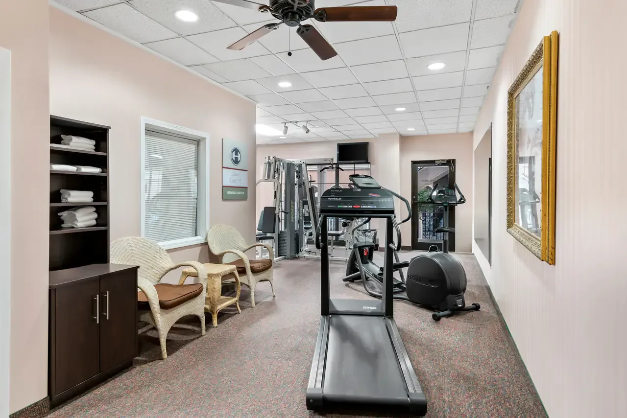 Indoor fitness center with treadmill in foreground, exercise machines in the background, chairs on the left, mirrors and picture frames on walls.