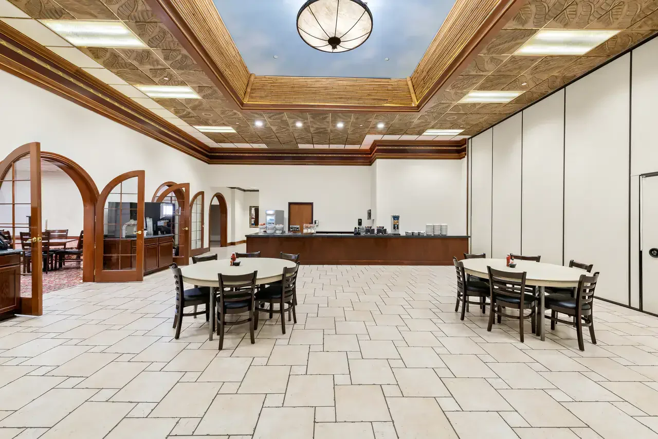 Empty banquet hall with round tables and chairs, a long side counter, open doorways, and decorative ceiling (likely part of a hotel or conference center).