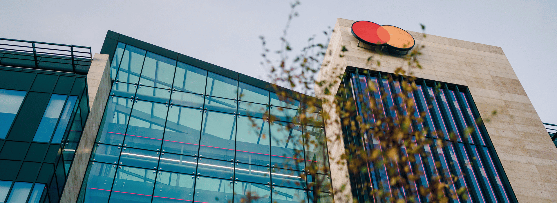 Modern building with glass windows and a beige stone facade, featuring a red and orange logo on the top right corner, and some blurred branches with leaves in the foreground.