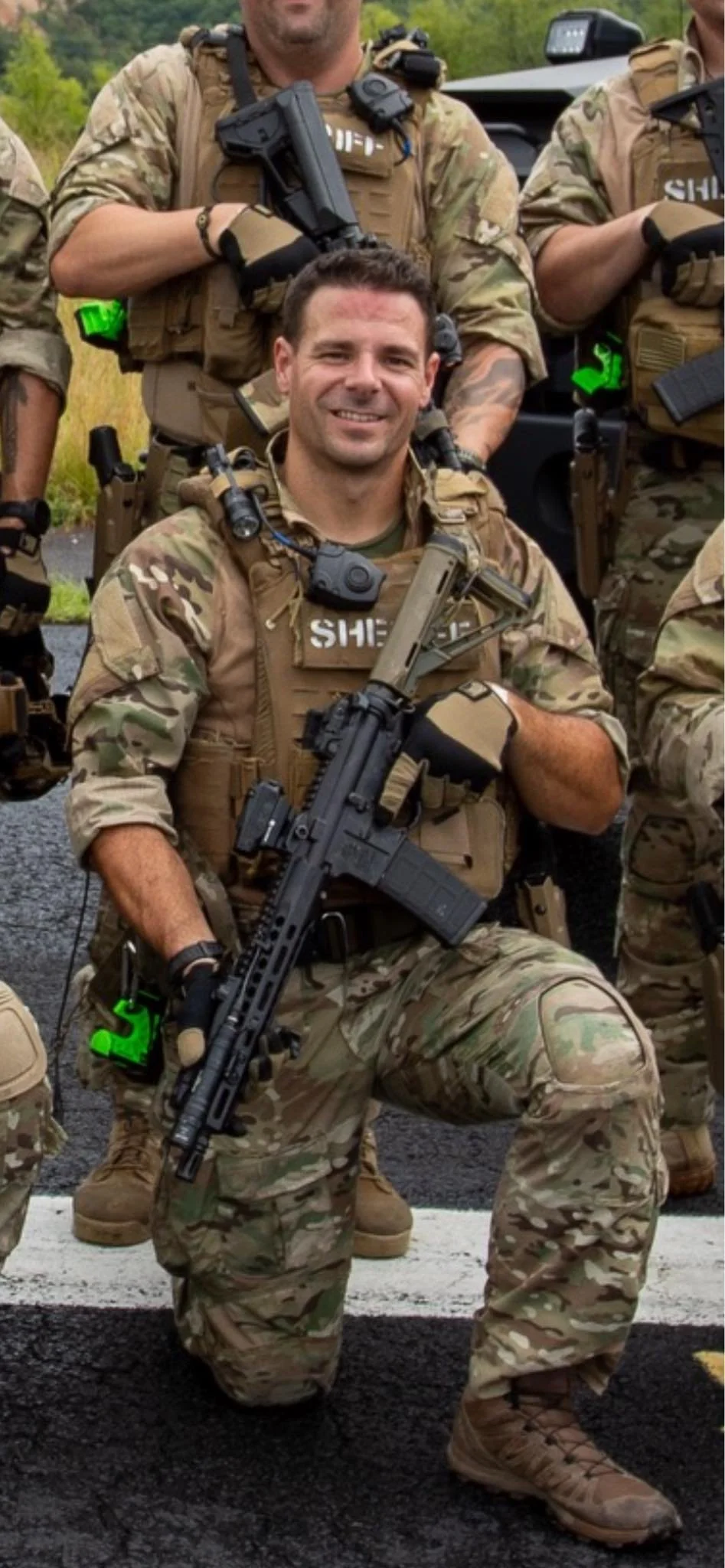 Kneeling soldier in camouflage uniform holding a rifle, with other soldiers standing behind him, all in military gear and uniforms.