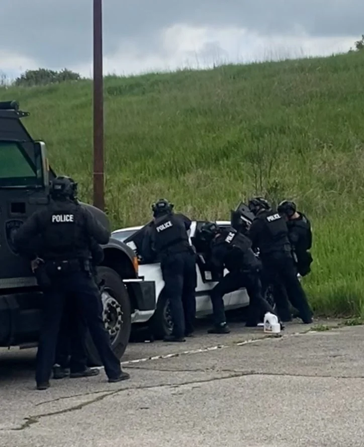 Five police officers wearing tactical gear are surrounding and inspecting the interior of a police car on the side of a rural road, with a grassy hill in the background.