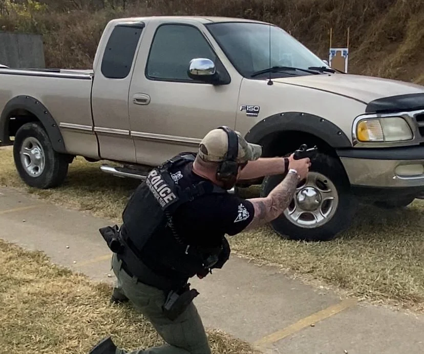 A police officer in tactical gear aiming a firearm while kneeling on the sidewalk near a beige pickup truck.