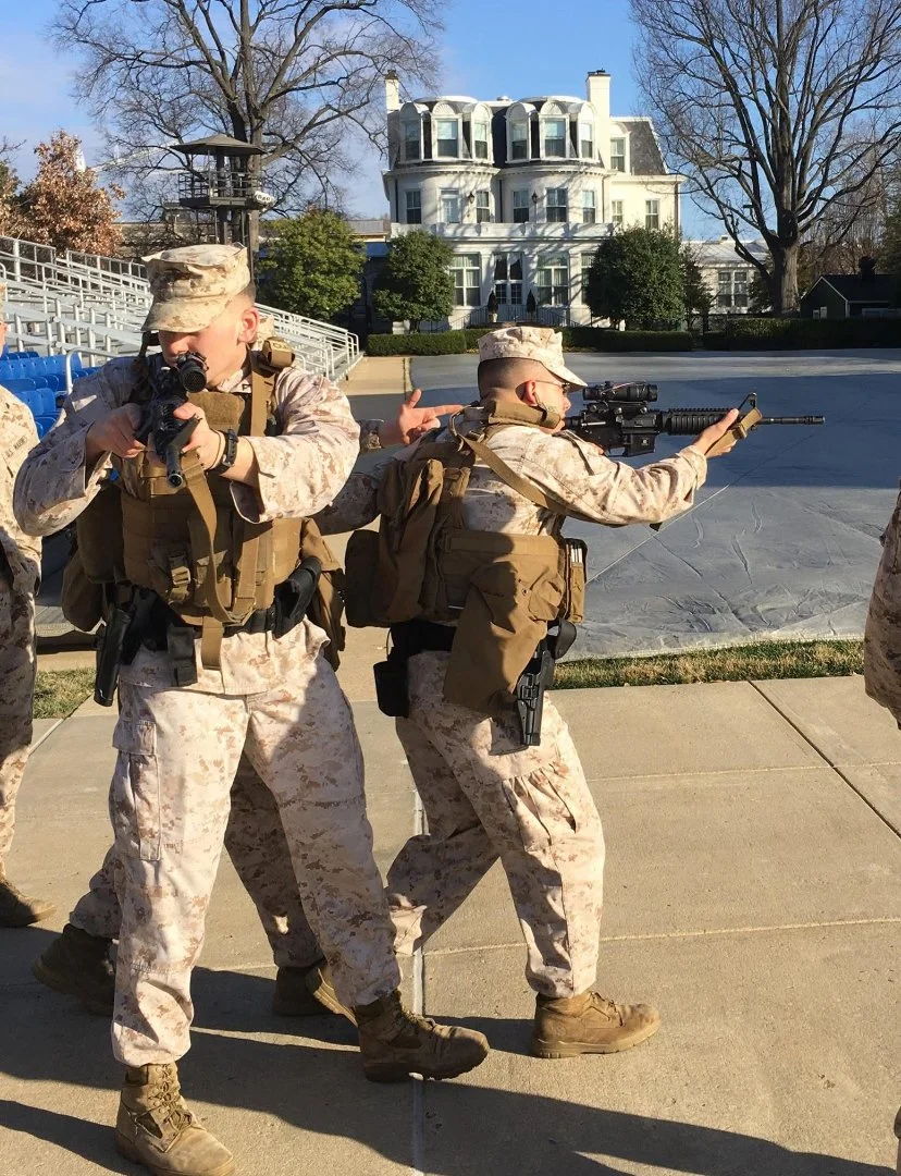 Two soldiers in camouflage uniforms aiming rifles, training or practicing on a sidewalk with a large white house and leafless trees in the background.